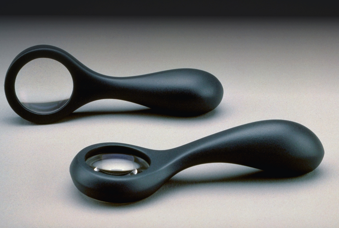 Two views of a black magnifying glass with soft, curved edges, propped on its side and lying on its back side