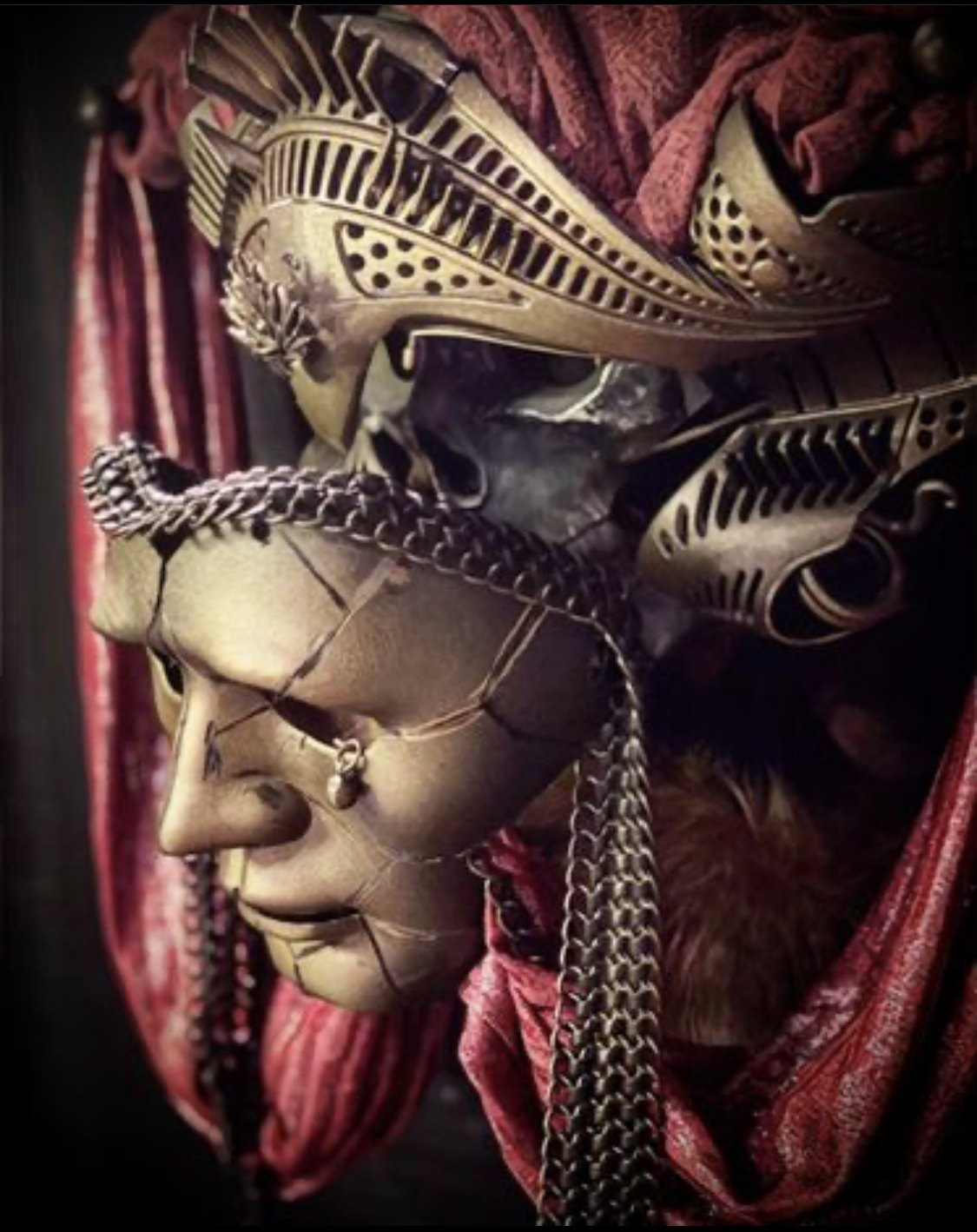 A mask of a human face made of pieced-together fragments, chain mail, and assorted components with circular and rectangular vents, draped in red brocade fabric