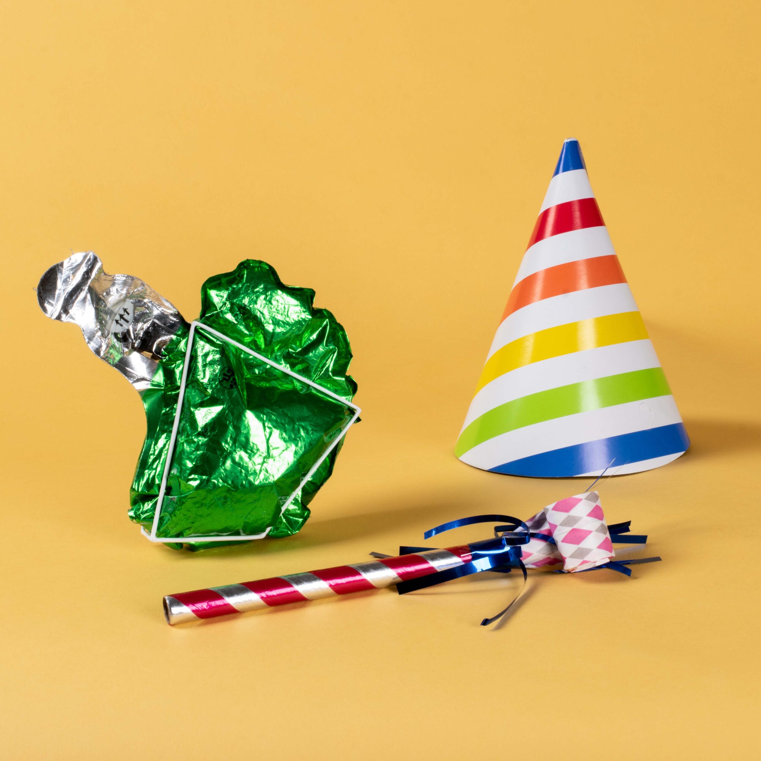 a striped party hat, a green foil and a party horn against a yellow background.