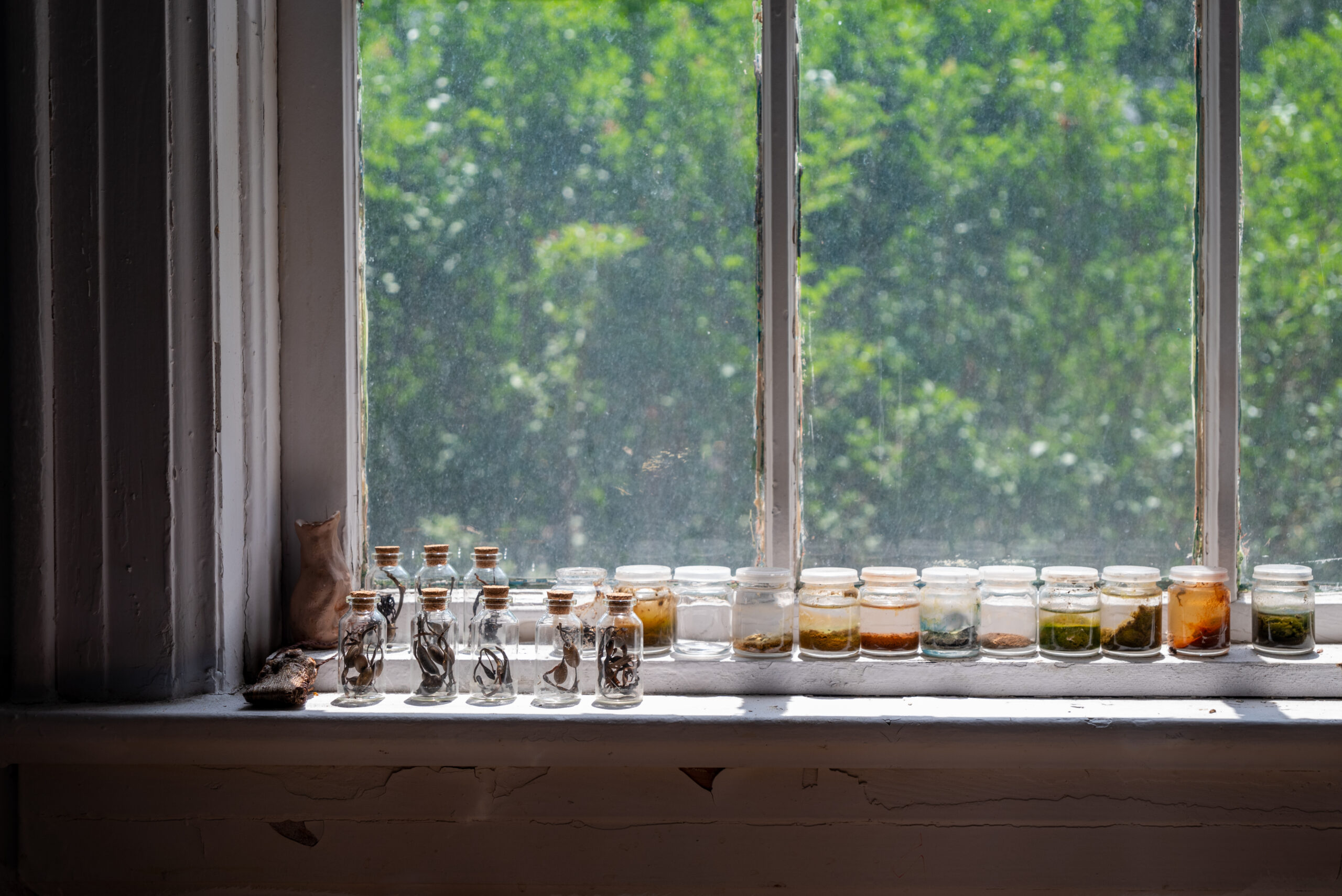Small glass jars with cork stoppers, filled with dried plants and colored powders, line a windowsill