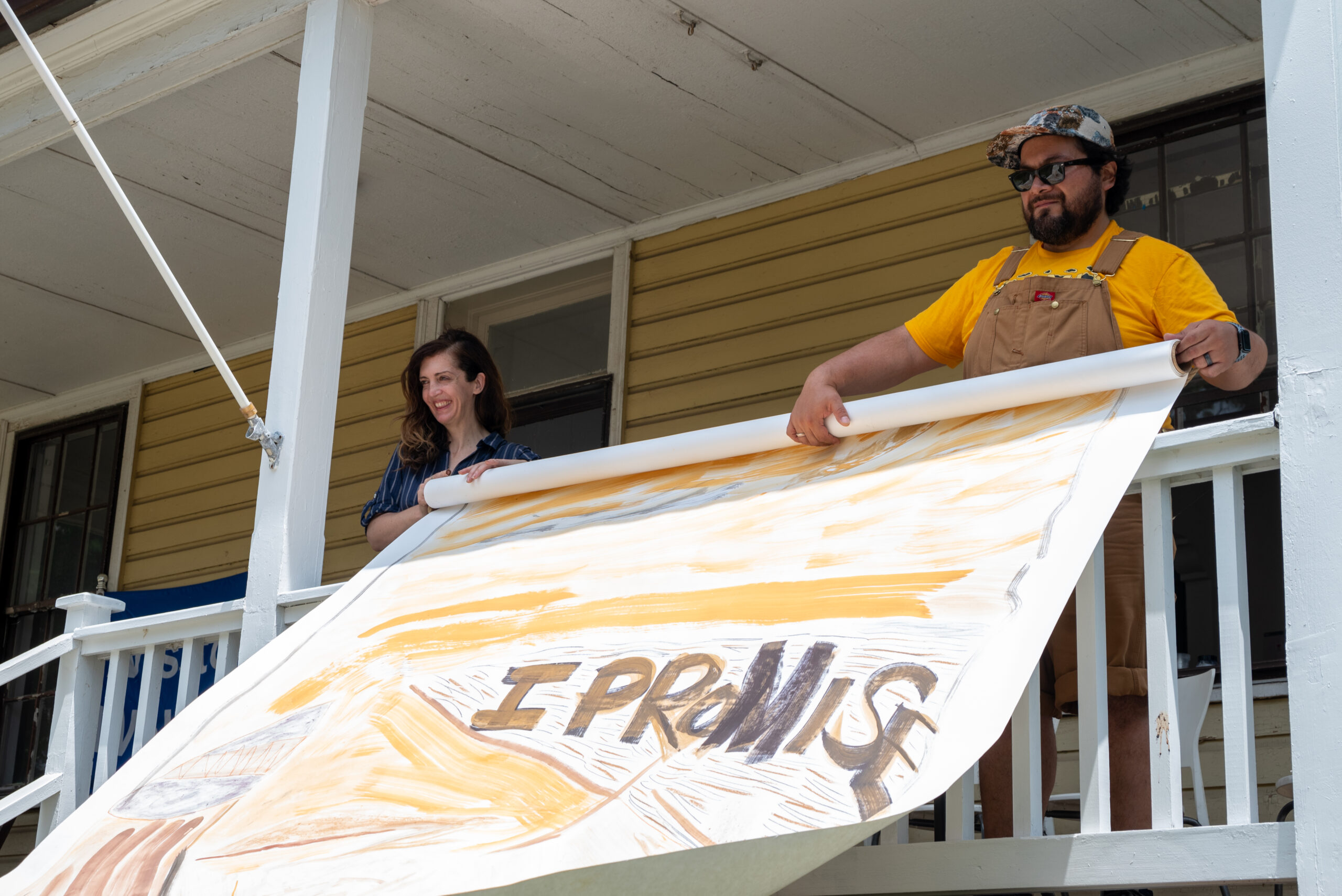 Two people stand on a porch holding a wide roll of paper painted in earthy tones that reads I Promise in wide capital letters