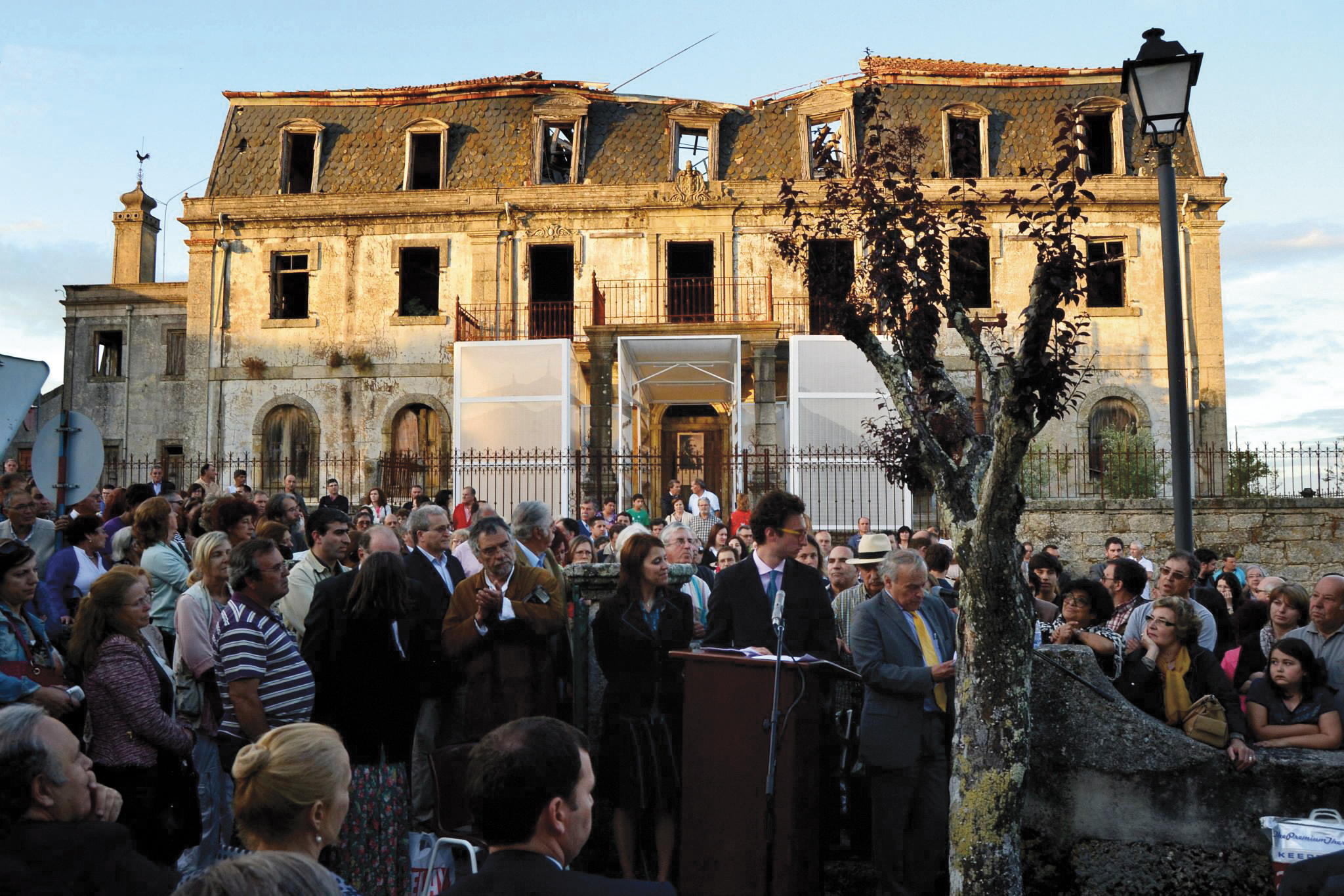 People gathered in front of a large stone home that has fallen into disrepair with translucent rectangular installation around the entryway