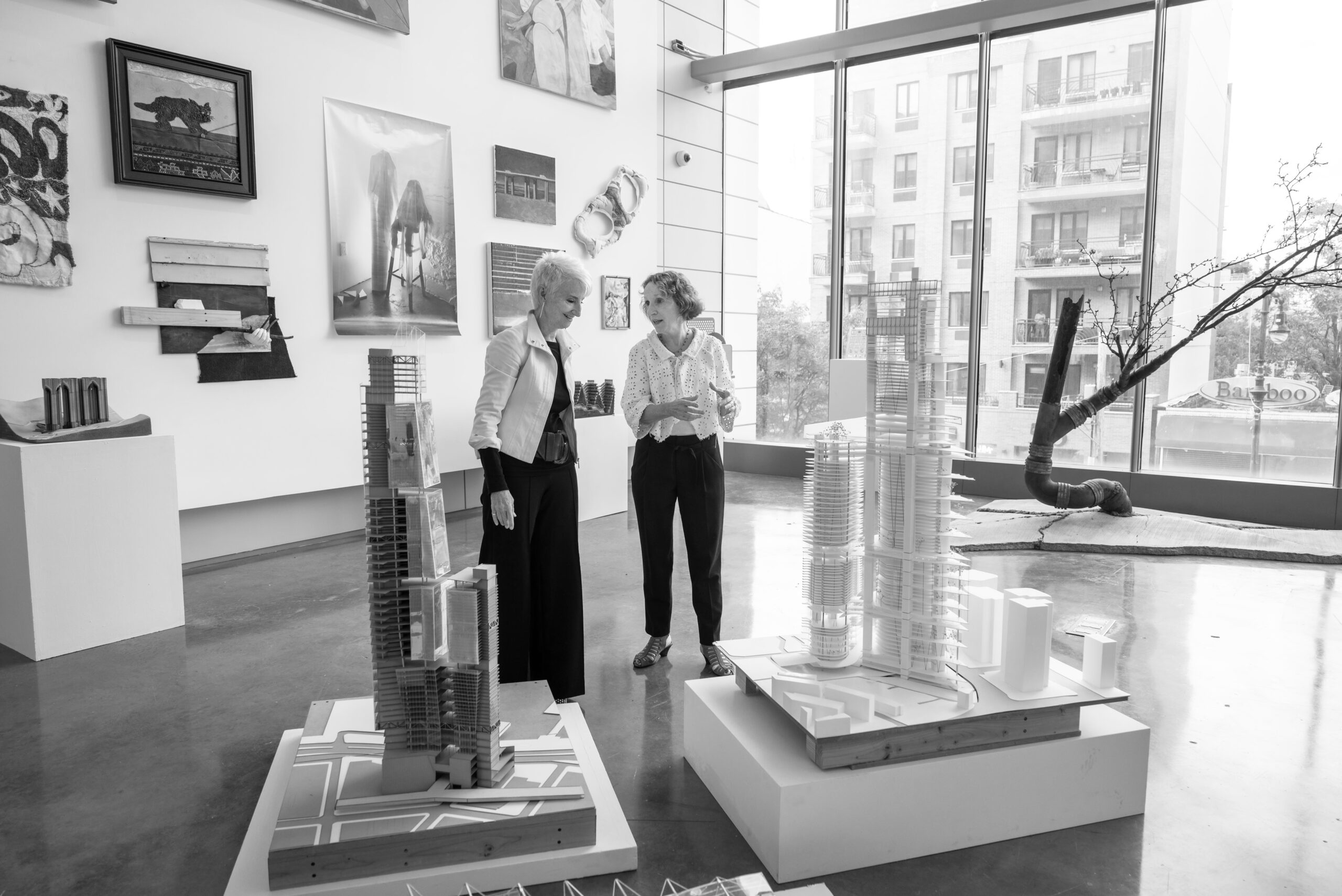 Frances Bronet and Donna Heiland gesture at architectural models in a gallery hung with artwork