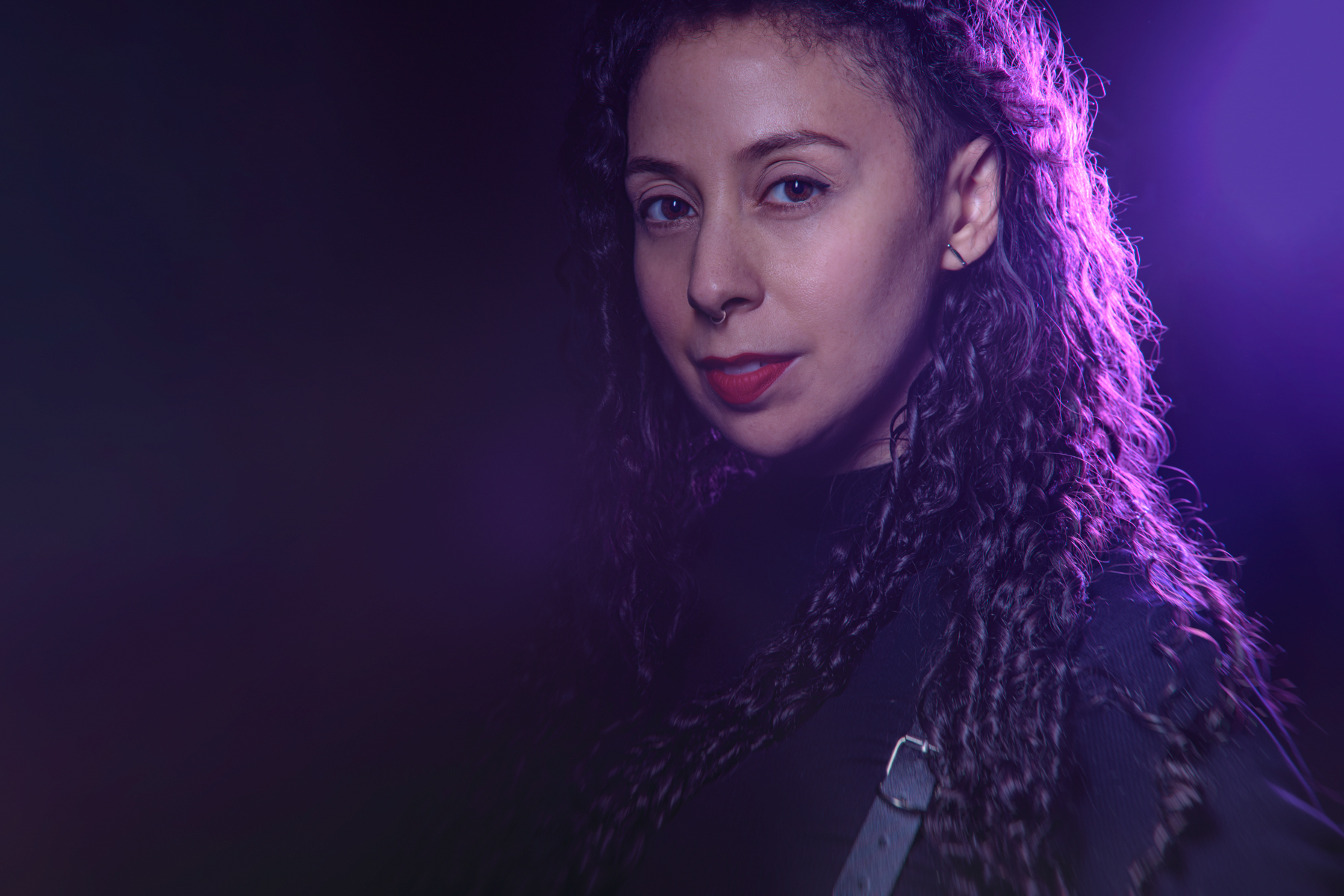 Nasreen Alkhateeb illuminated in purple light, with long, curly brown hair pulled back, brown eyes looking at the photographer’s lens, black turtleneck, and a gold hoop septum ring