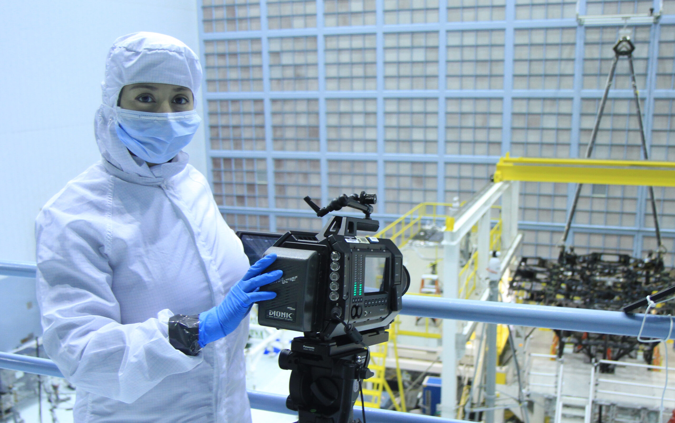 Nasreen Alkhateeb in white protective suit, gloves, and mask in a clean room with camera and Webb telescope assembly in background