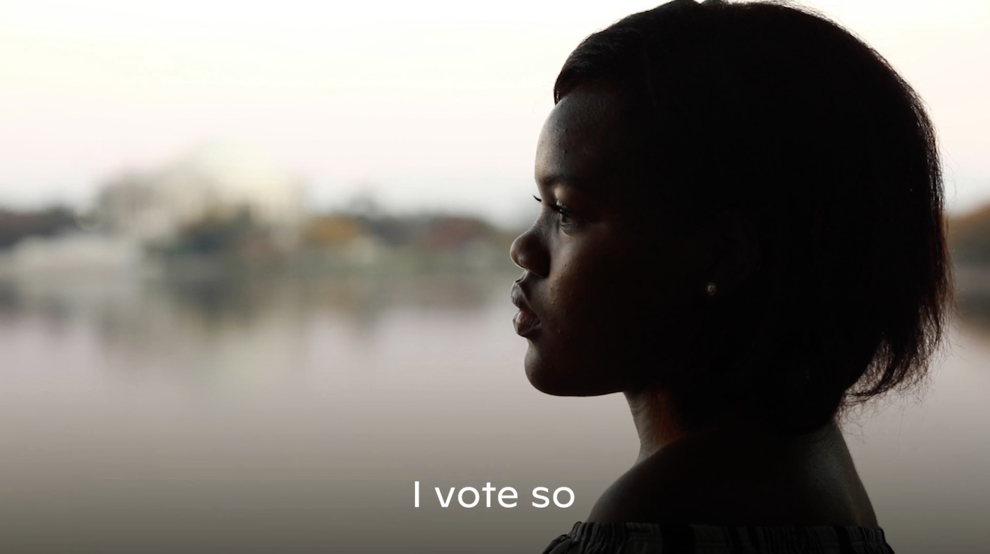 Person illuminated in gently light with dewy brown skin and cropped dark hair, in profile against a still body of water, with the words I vote so at the bottom of the image