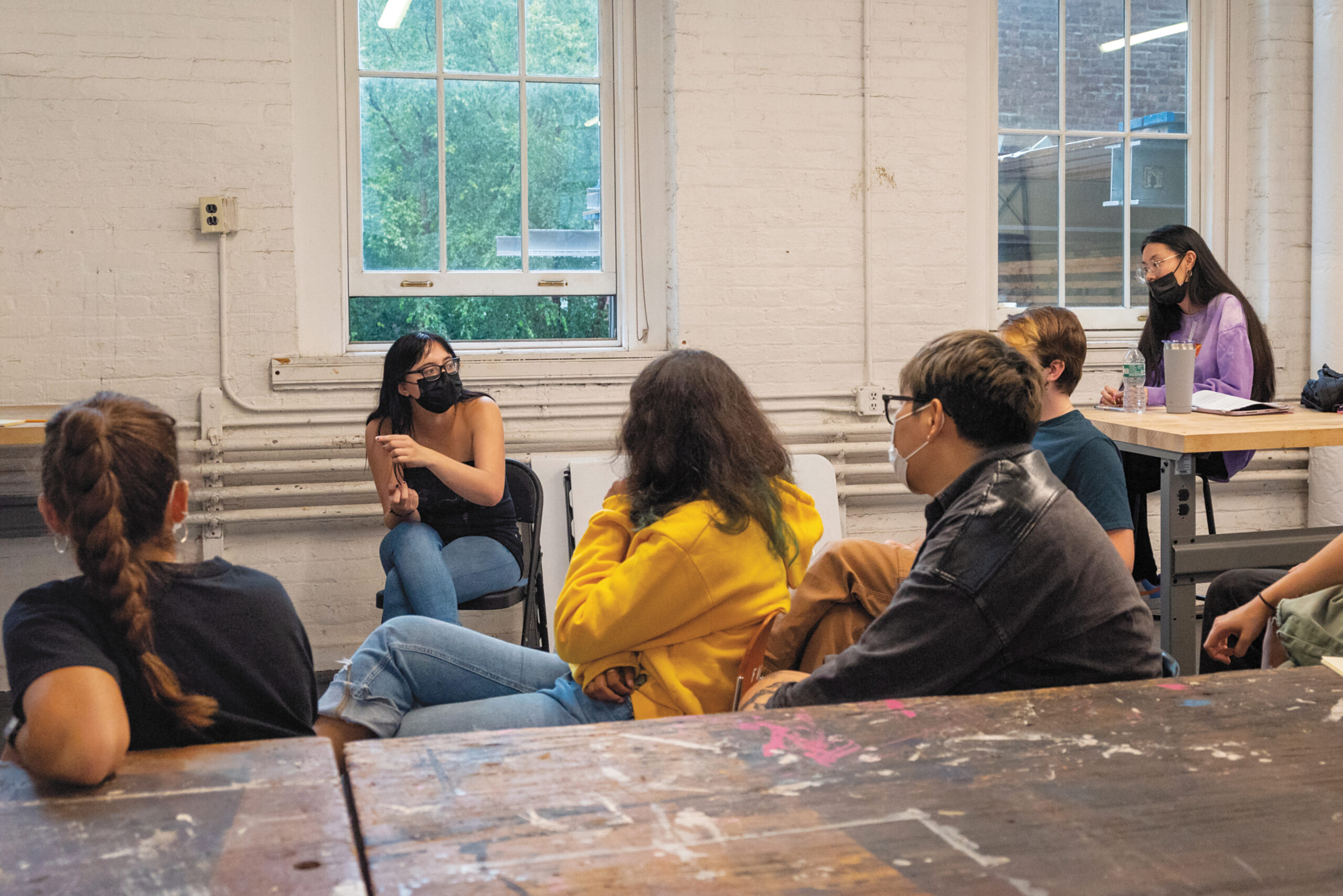 Six students sit in an arc in a white walled studio classroom gesturing in a discussion of an unseen work
