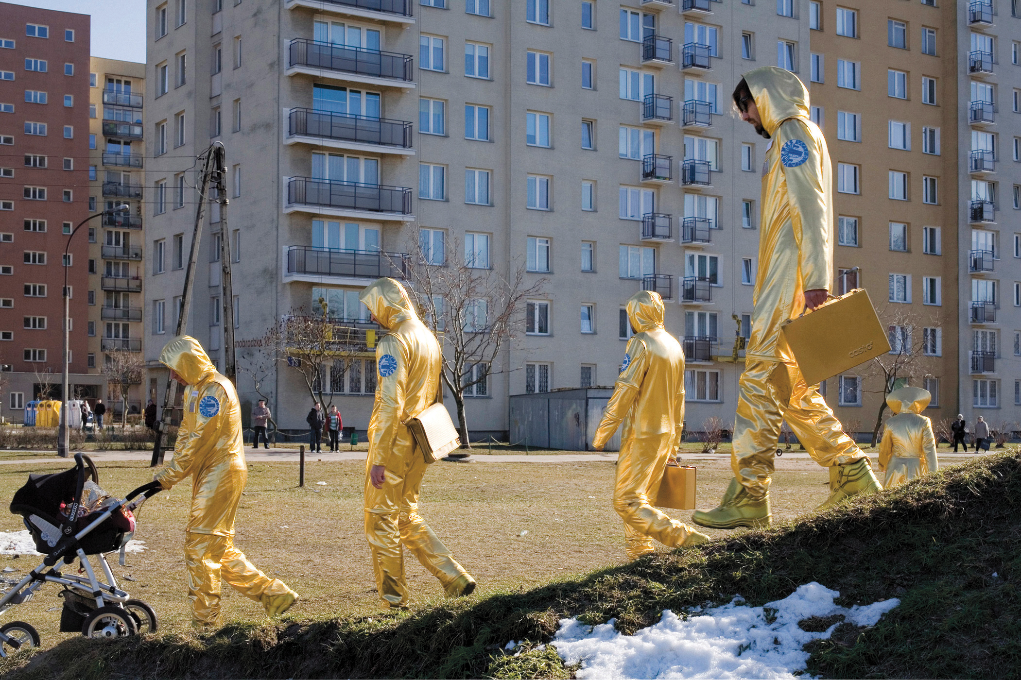 People in gold space suits walk over a hill in front of a cluster of residential towers, carrying briefcases and pushing a baby carriage