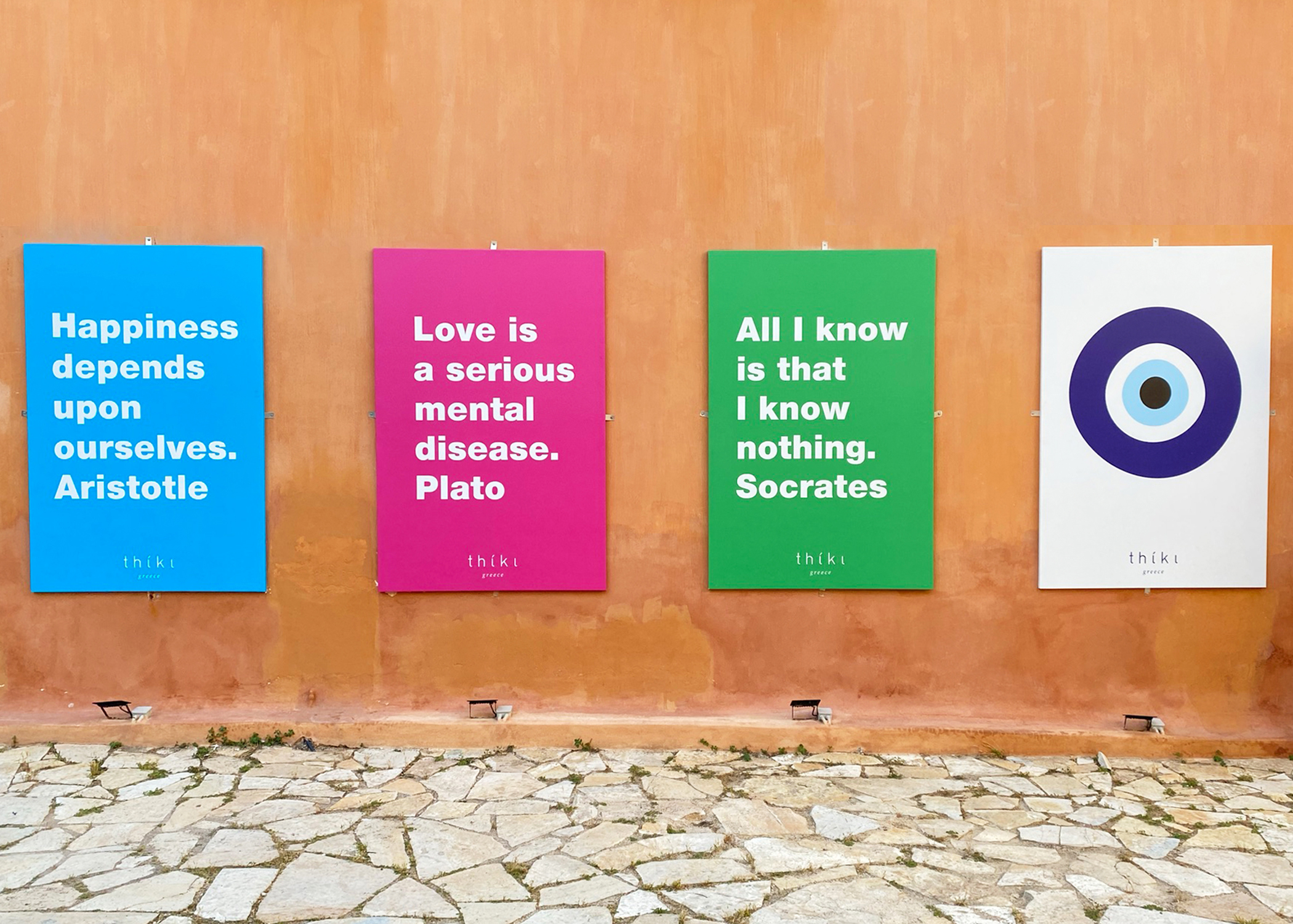 Four signs hang on a clay-colored exterior wall, with quotes from Aristotle, Socrates, and Plato and an illustration of the evil eye