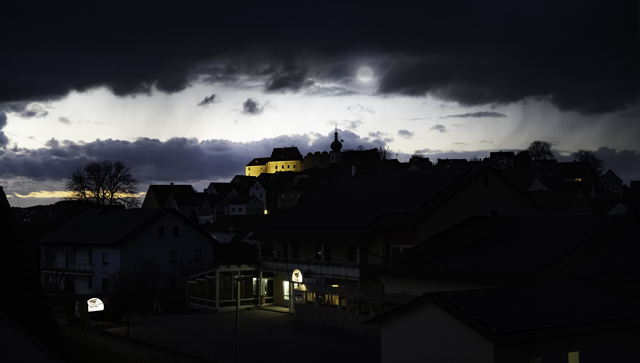 A cluster of houses and scattered bare-limbed trees on a small hillside beneath a dimly lit sky and dark clouds