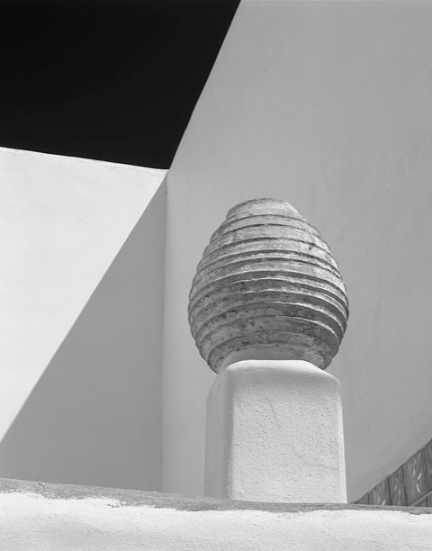 Black-and-white photo of a round, ridged vessel placed before angled walls of a building that cast sharp shadows