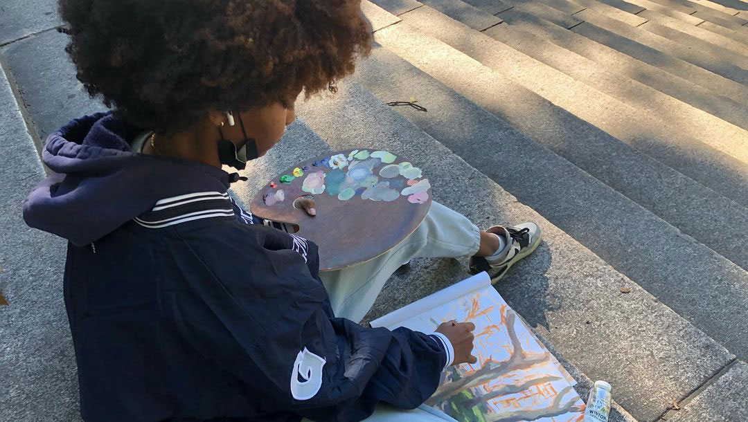 A person with a painting palette in one hand and painting on a sketchbook with the other