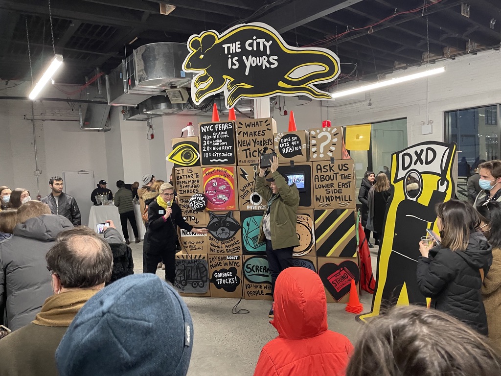 people gathered in a gallery, circled around the New York Alive installation, with one person on a microphone demonstrating with another person. Installation is made of paper and cardboard, with hand written signs and art in a boxed grid, with a large cut-out of a rat on the top-- with the words 
