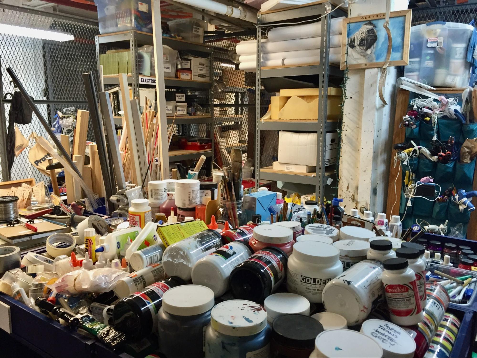 a photo of a supply closet, with a large amount of paint and paint products in the foreground, with grey metal shelving in the background containing boxes, canvases, and rolled up easels