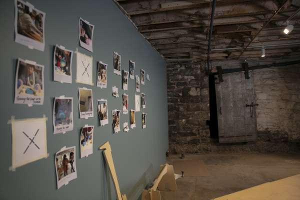A photo depicting a studio space with exposed brick and rafters on the ceiling, with a sliding metal door on one wall, the opposite wall is made of smooth drywall, with about 20 photographs with captions under them, some with writing on the pictures