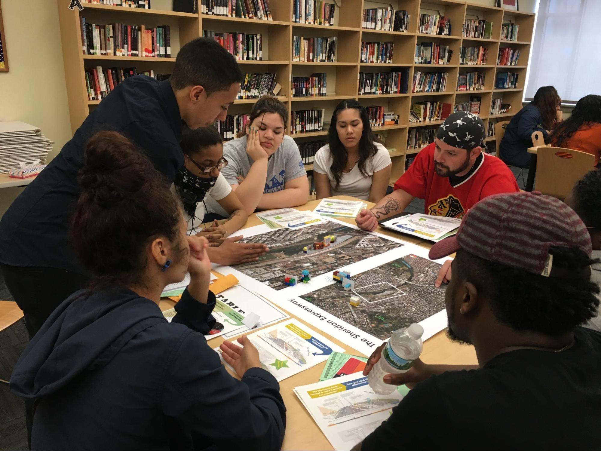 Students gathered in a library, seated around a table, with one student standing-- working with a professor seated across the table