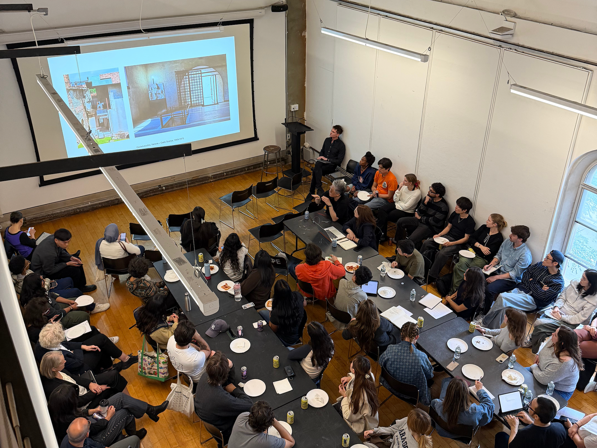 An overhead view of a classroom or lecture room showing a group of students and faculty seated around long tables, attentively watching a presentation projected on a large screen at the front of the room. The presentation features architectural images labeled “Corbusier, Venice Case Study 1953–1973.” Attendees have notebooks, laptops, and plates of food in front of them, indicating an informal yet engaged academic discussion.