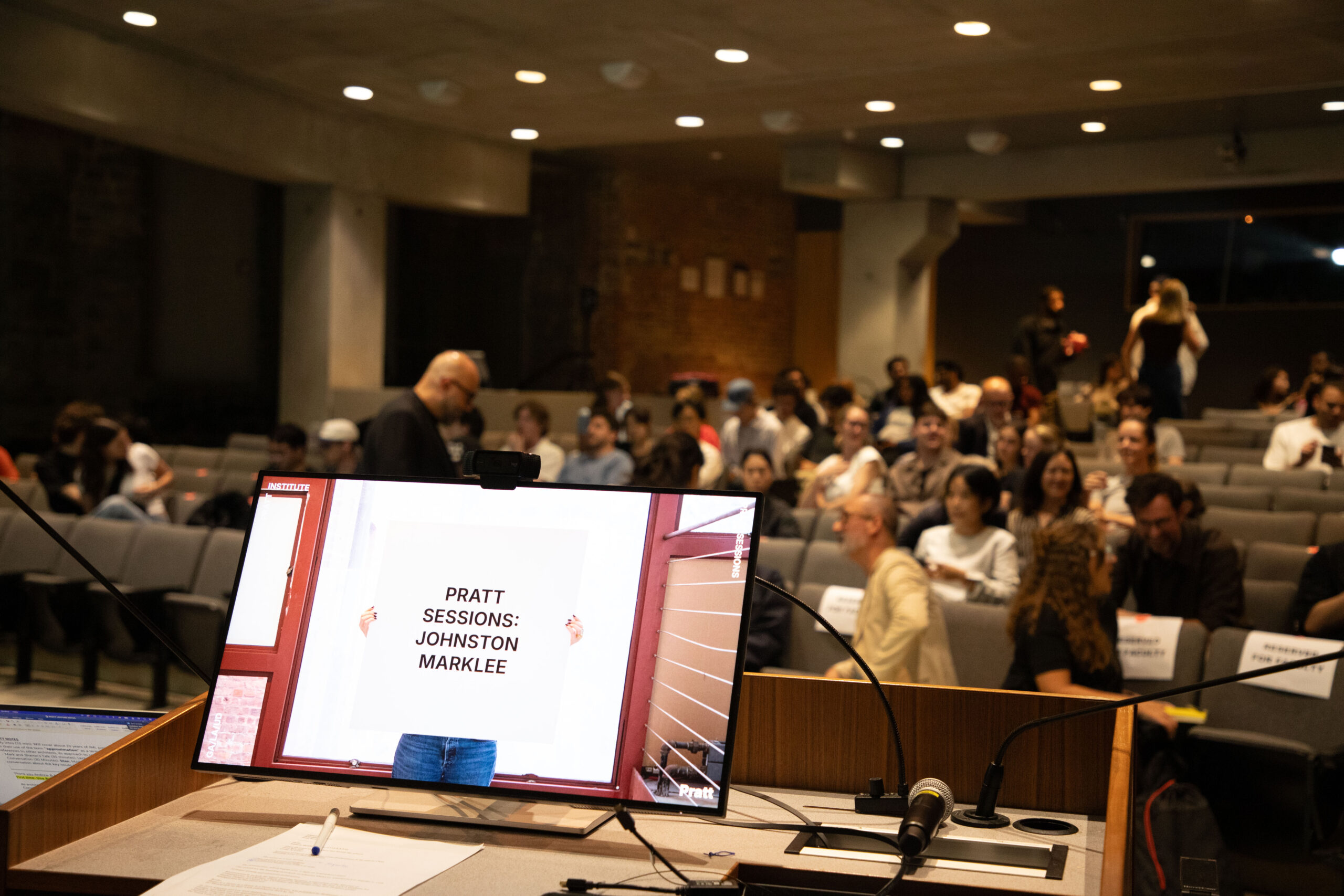 Four speakers sit in red chairs on stage at a Pratt Institute auditorium, engaging in a panel discussion in front of a large projected aerial image of a contemporary industrial complex. Audience members sit in rows facing the stage.