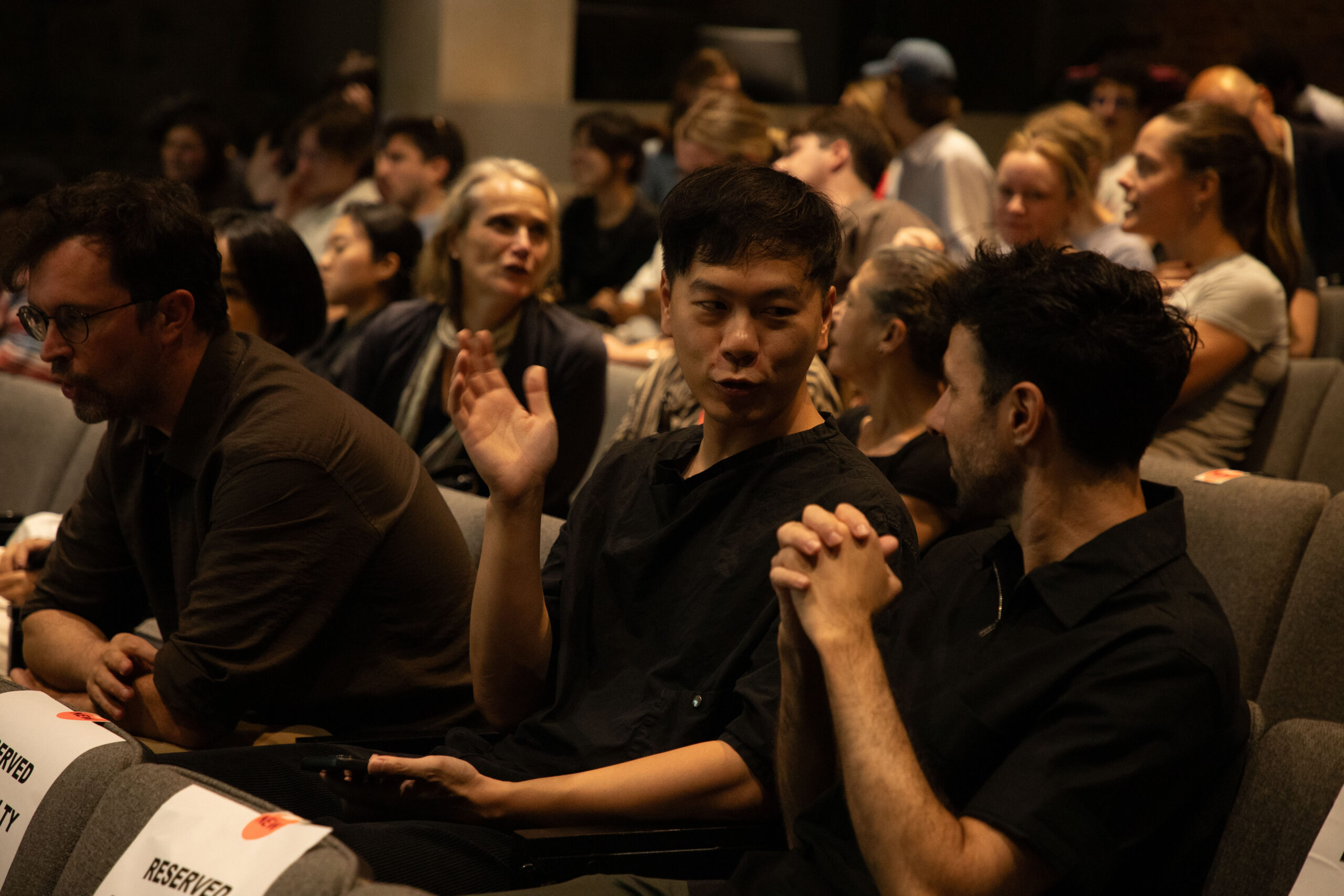 A close-up view from behind a lectern shows a monitor displaying the text “Pratt Sessions: Johnston Marklee” as audience members take their seats in an auditorium before the event begins.