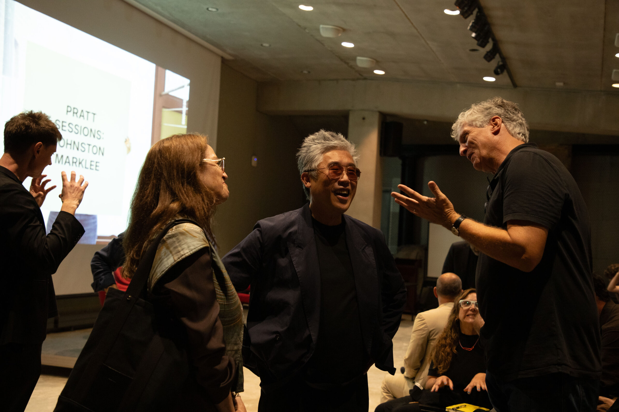 A man wearing glasses speaks to another attendee in conversation at a Pratt Institute event, with people mingling in the background under soft lighting.