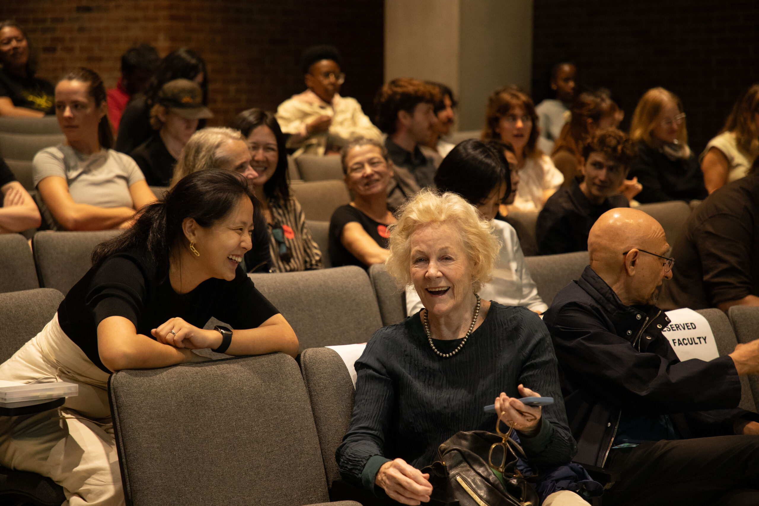 Audience members seated in gray auditorium chairs engage in lively conversation before a talk begins, with several seats labeled “Reserved Faculty.”