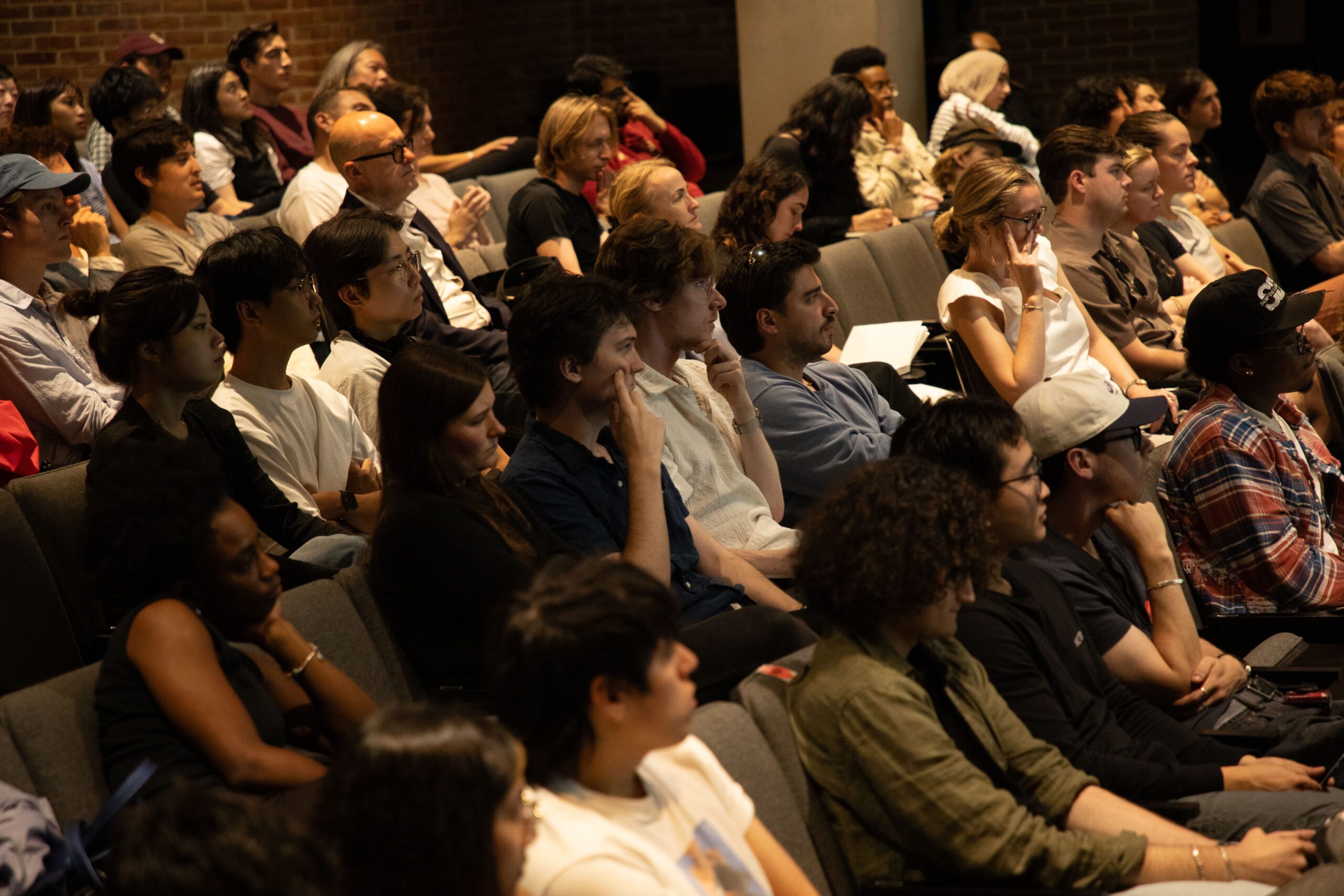 Two people converse animatedly in the foreground while another group mingles nearby in an auditorium setting. A large screen behind them reads “Pratt Sessions: Johnston Marklee.”