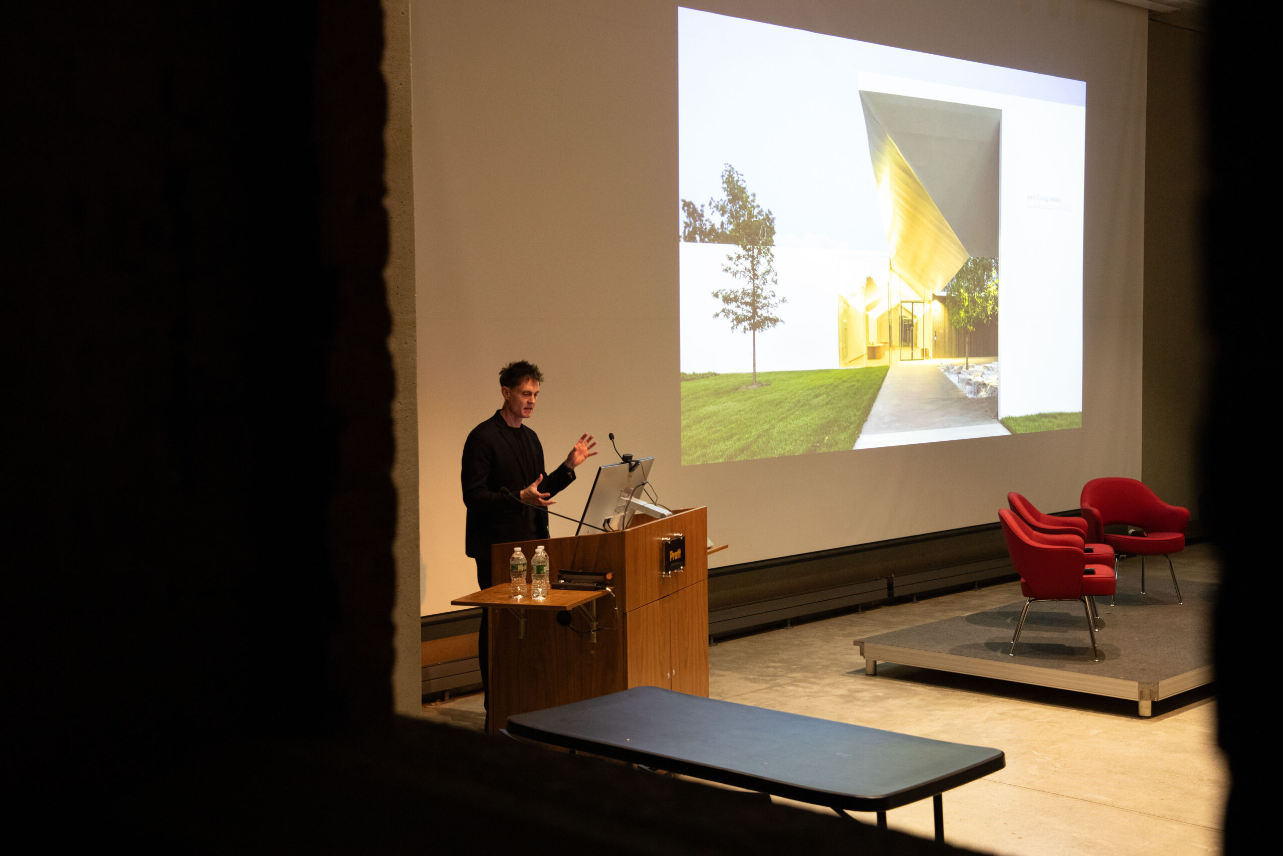 Two women seated in the audience share a laugh before an event at Pratt Institute, surrounded by other attendees chatting and waiting for the session to start.