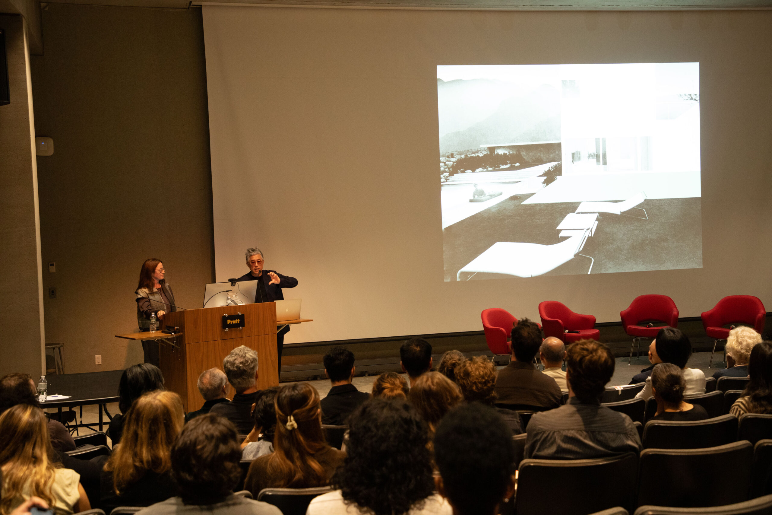 Audience members in a lecture hall focus intently on a presentation projected at the front, sitting in rows of gray seats under warm lighting.