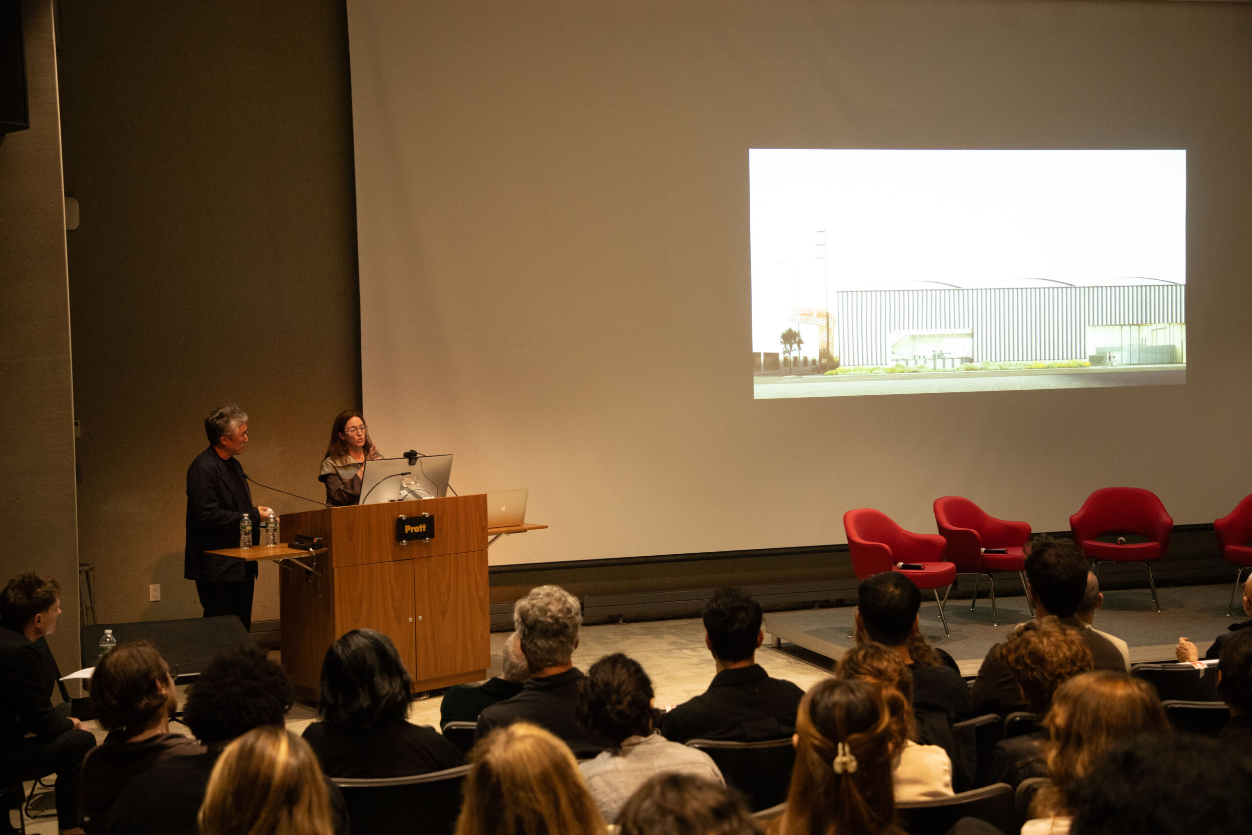 Two presenters stand at a wooden podium in front of a large projected black-and-white image of a modernist outdoor space with lounge chairs, speaking to an attentive audience in a Pratt auditorium.