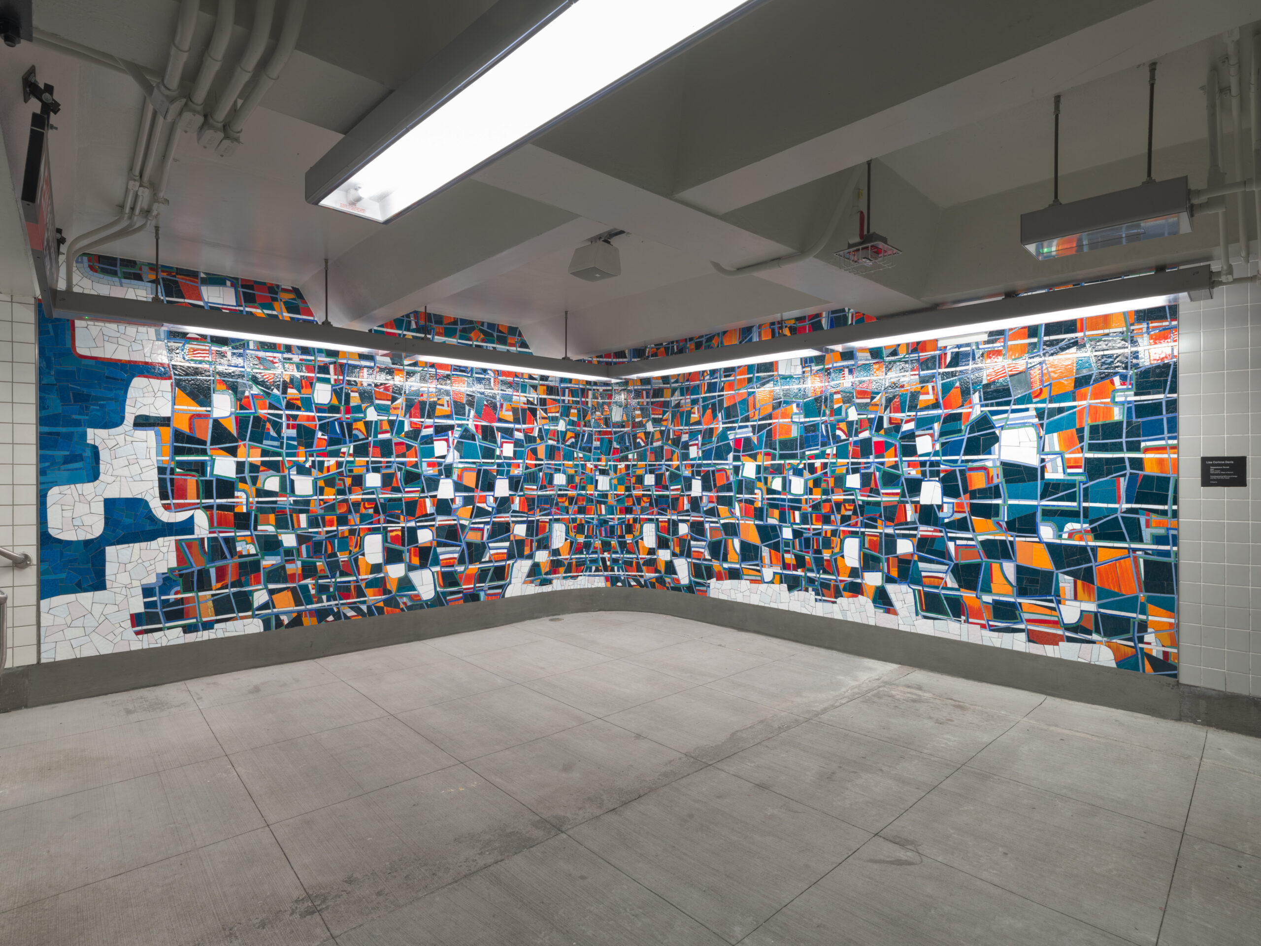 A large abstract mosaic mural inside the 68th Street–Hunter College subway station in New York City. The artwork covers two walls of a corner, composed of irregular geometric shapes in deep blues, bright oranges, reds, and whites, arranged like fragmented tiles. The mural creates a dynamic, puzzle-like visual effect under bright fluorescent lighting in the underground station.