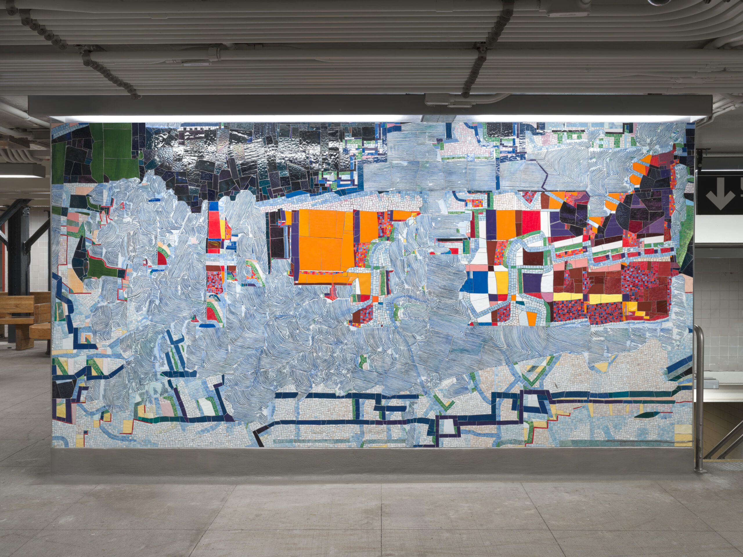 Abstract mosaic mural at the 68th Street–Hunter College subway station in New York City. The piece combines bold blocks of color—red, orange, yellow, and blue—with textured gray and white patterns resembling brushstrokes. The mural stretches across a flat wall, incorporating both dense geometric areas and open sections, evoking a layered, map-like composition. It is installed in a brightly lit transit concourse with tiled walls and exposed ceiling pipes.
