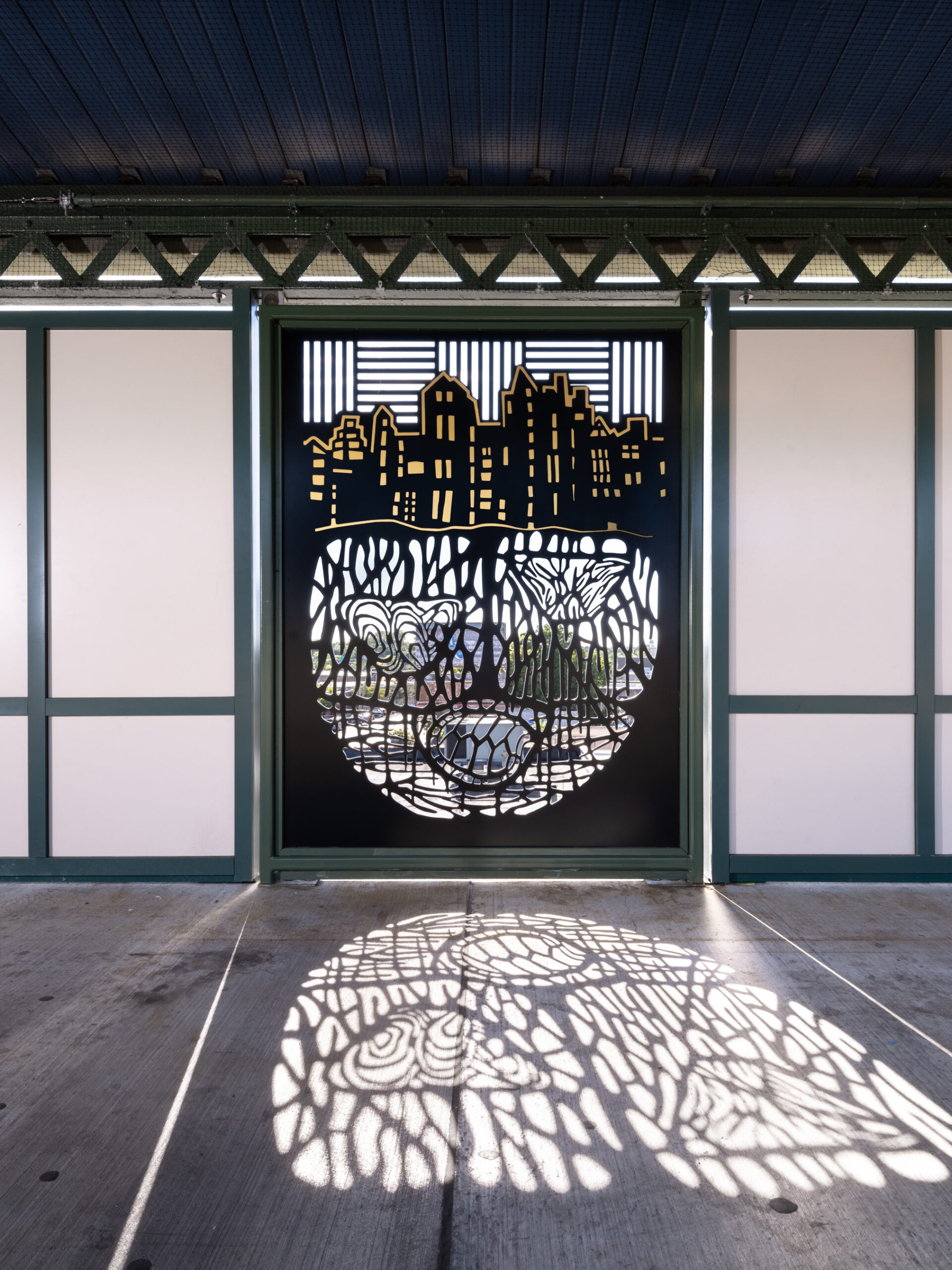 Art installation at the Westchester Square subway station. A decorative metal panel depicts a city skyline above a pattern of organic shapes, with light filtering through to cast intricate shadows on the station floor. The cut-out design creates a circular motif combining urban and natural imagery, adding visual interest and depth to the station environment.