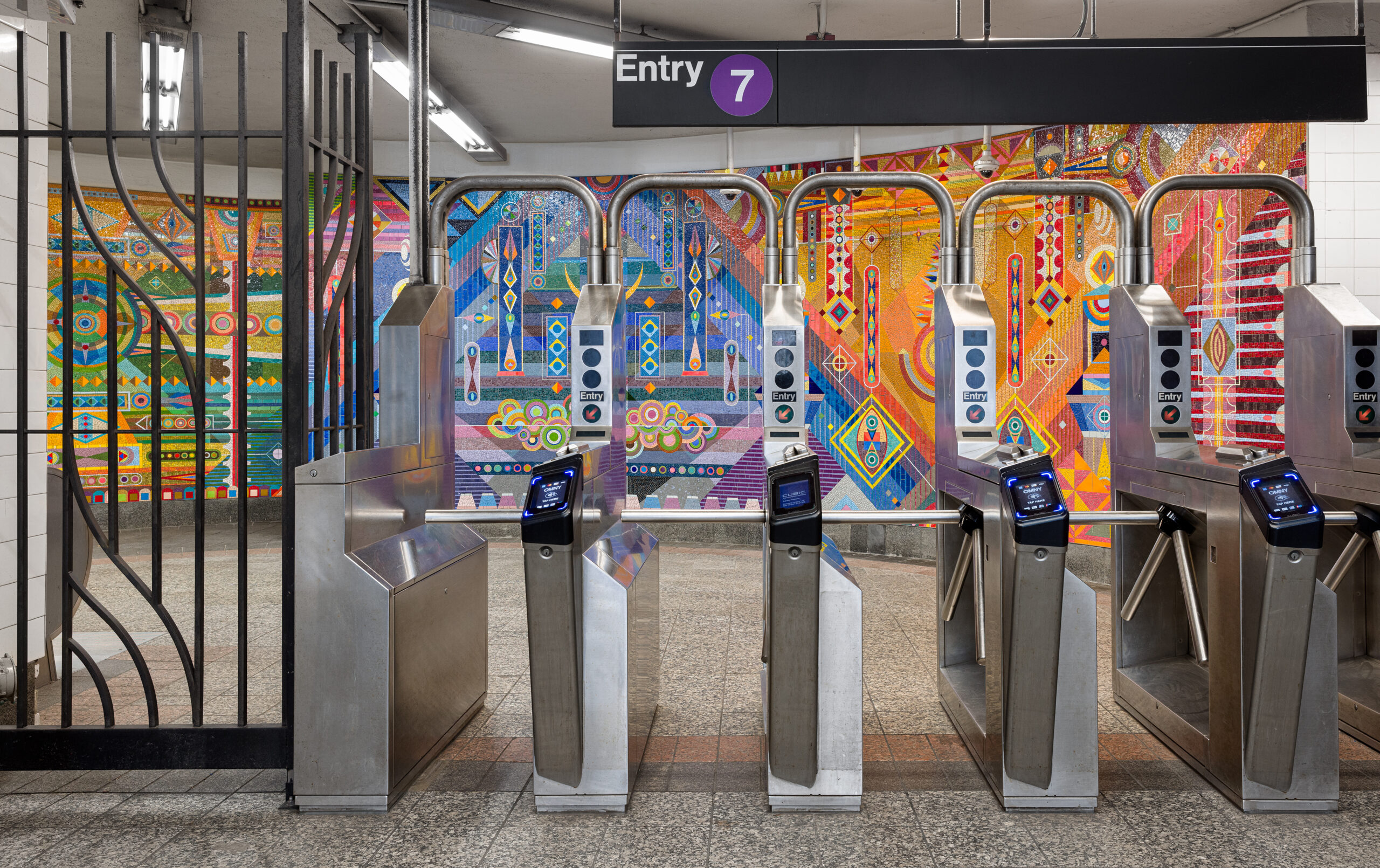 Interior view of a New York City subway entrance for the 7 line. Stainless steel turnstiles with contactless payment readers face a large, colorful mosaic mural covering the wall behind them. The mural features abstract geometric shapes and patterns in vibrant shades of orange, red, blue, and green, creating a lively and artistic atmosphere in the transit space.