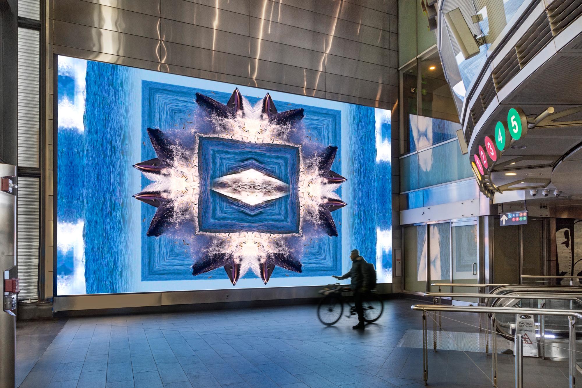 A large digital screen displays an abstract image featuring shades of blue and geometric shapes, set in a modern indoor environment. A person riding a bicycle is moving past the screen, with colorful circular indicators visible on the nearby wall. The floor is tiled, and the overall space appears to be a transport hub or public area.