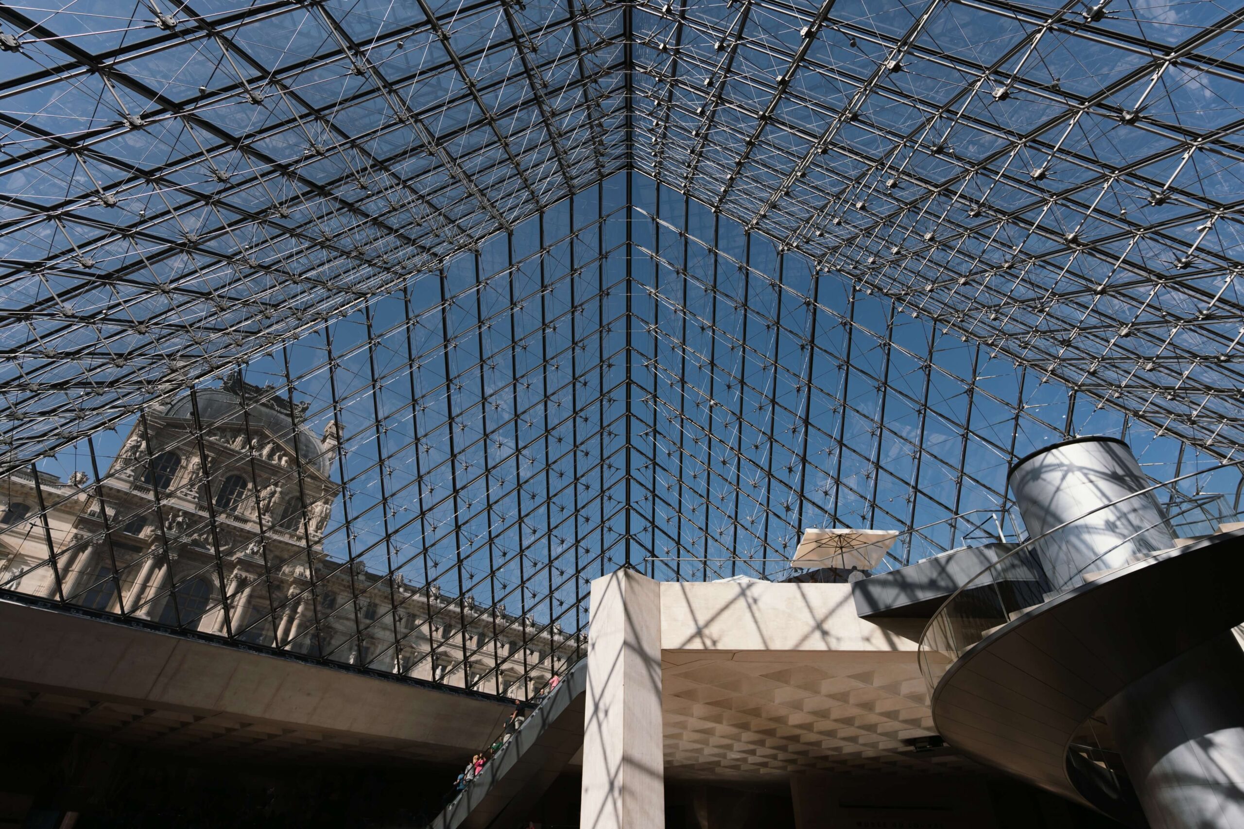 A panoramic phot of the underside of Louvre's glass pyramidal dome. A blue sky is visible through the glass.