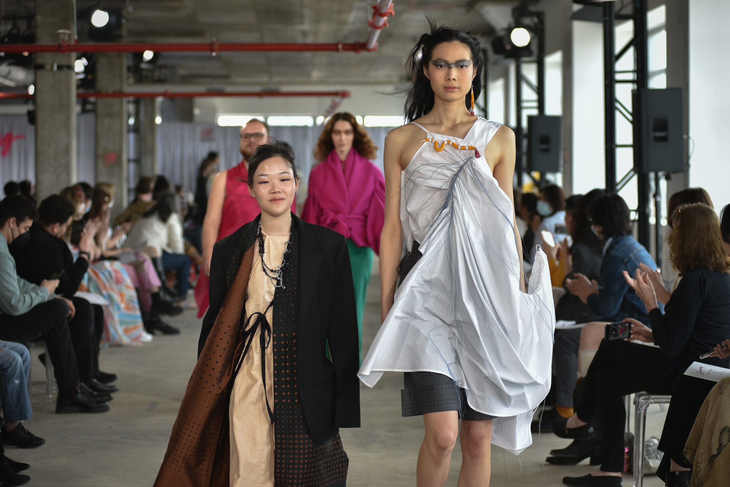 fashion show, held in concrete room with spot lighting fixtures, people seated in a rows on either side of the runway, with designer in foreground, walking alongside models in the show