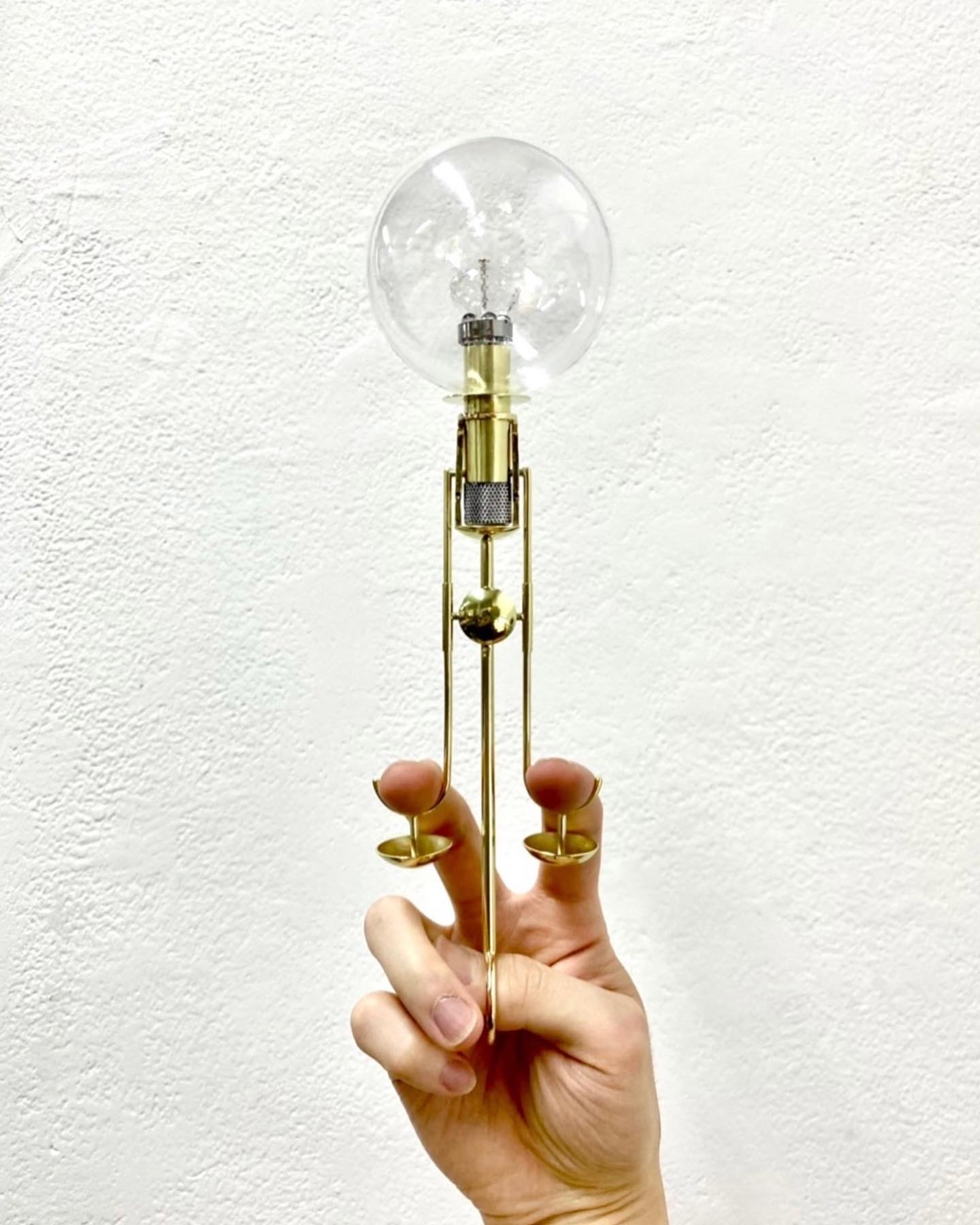 A hand holds gold-colored metal object in front of a white wall. The object has a large circular glass orb encompassing what appears to be either a light filament or an oil wick. This site atop a gold base which comes out of the glass orb and connects to three thin pieces of metal being held by the hand.