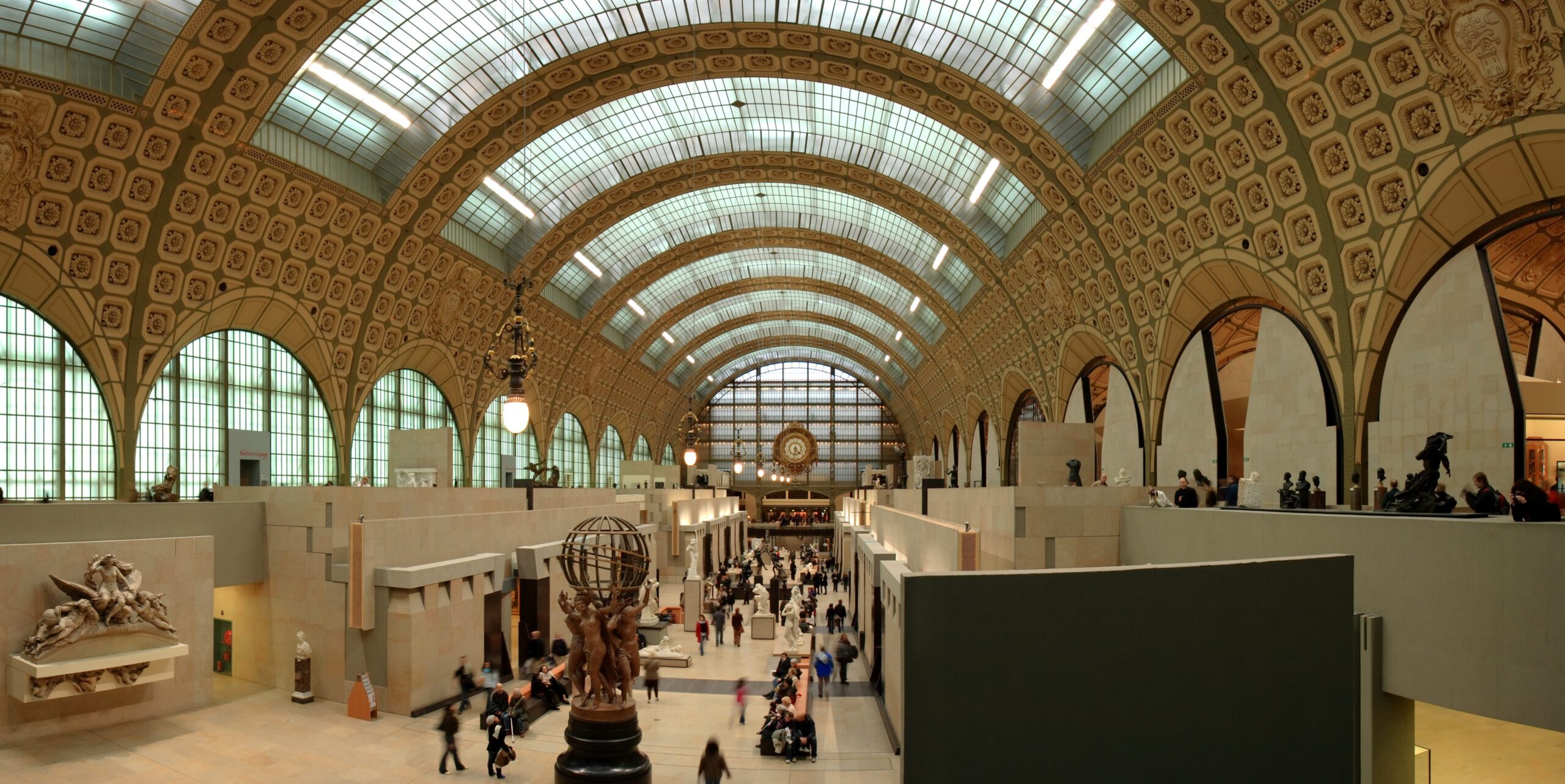 Panoramic image of the inside of a museum. There is a large arched ceiling ceiling and there is a stream of people of looking at exhibits scattered throughout.