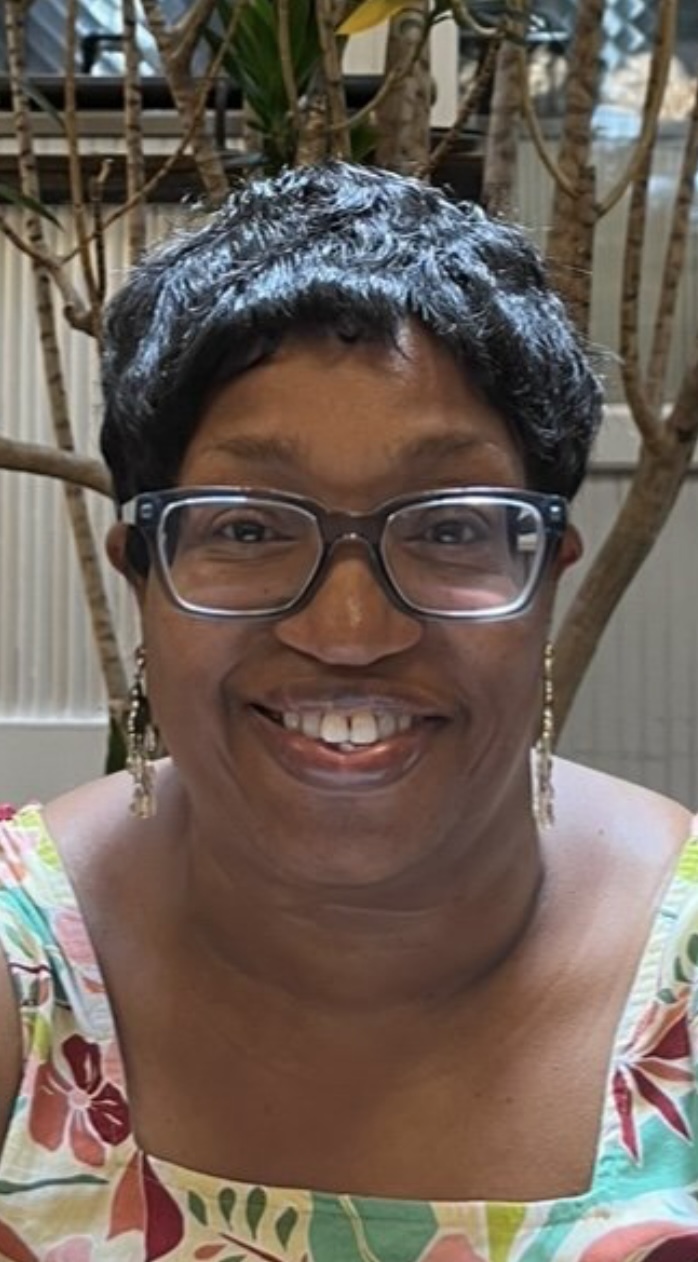 A woman with short black hair, glasses, and dangling earrings smiles at the camera. She is wearing a floral-patterned dress with shades of green, orange, and white. The background features potted plants and a greenhouse-like setting with natural lighting.
