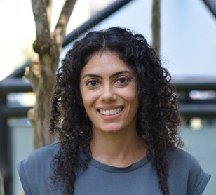 A woman with curly black hair smiles warmly at the camera. She wears a dark gray t-shirt. The background features tree branches and a metal structure, possibly in a park or an outdoor setting.