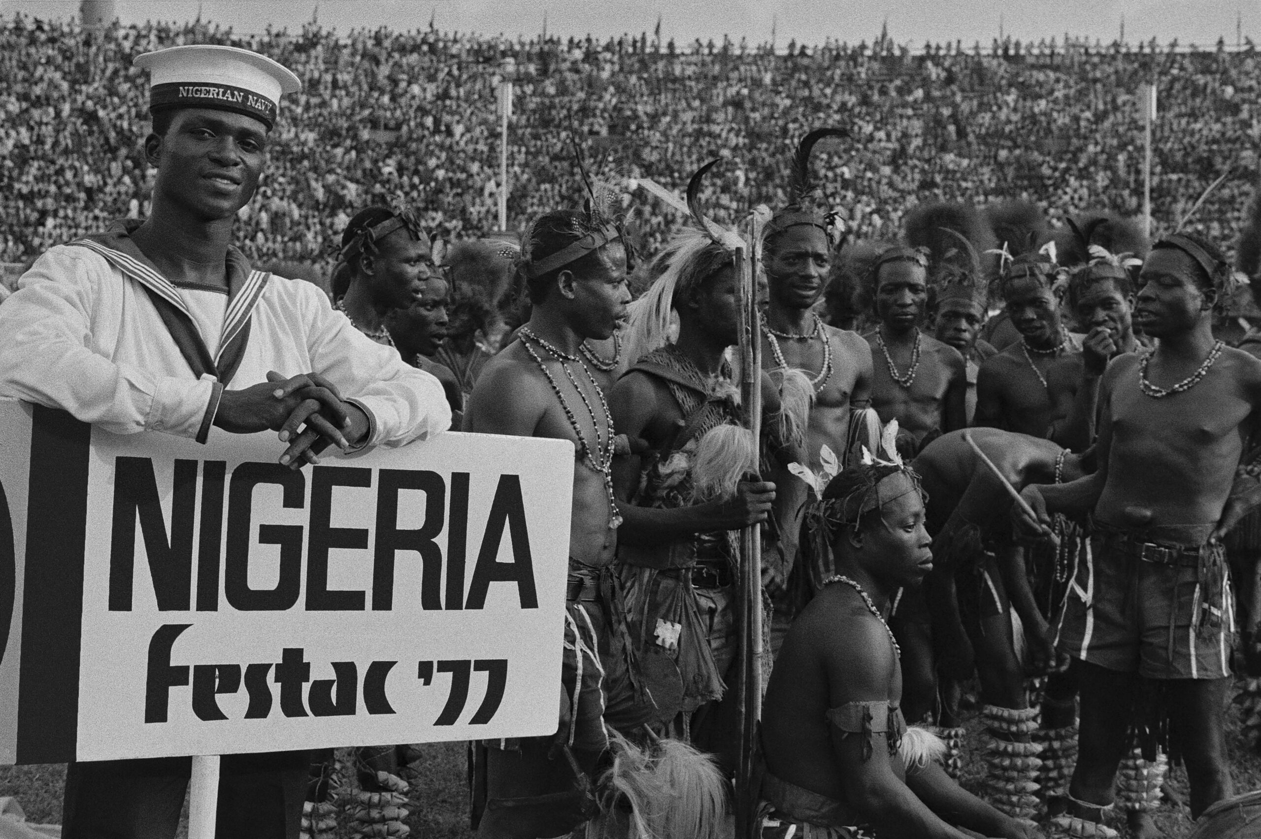 Person in sailor uniform poses with sign reading Nigeria Festac 77 in front of a stadium full of people from different African nations