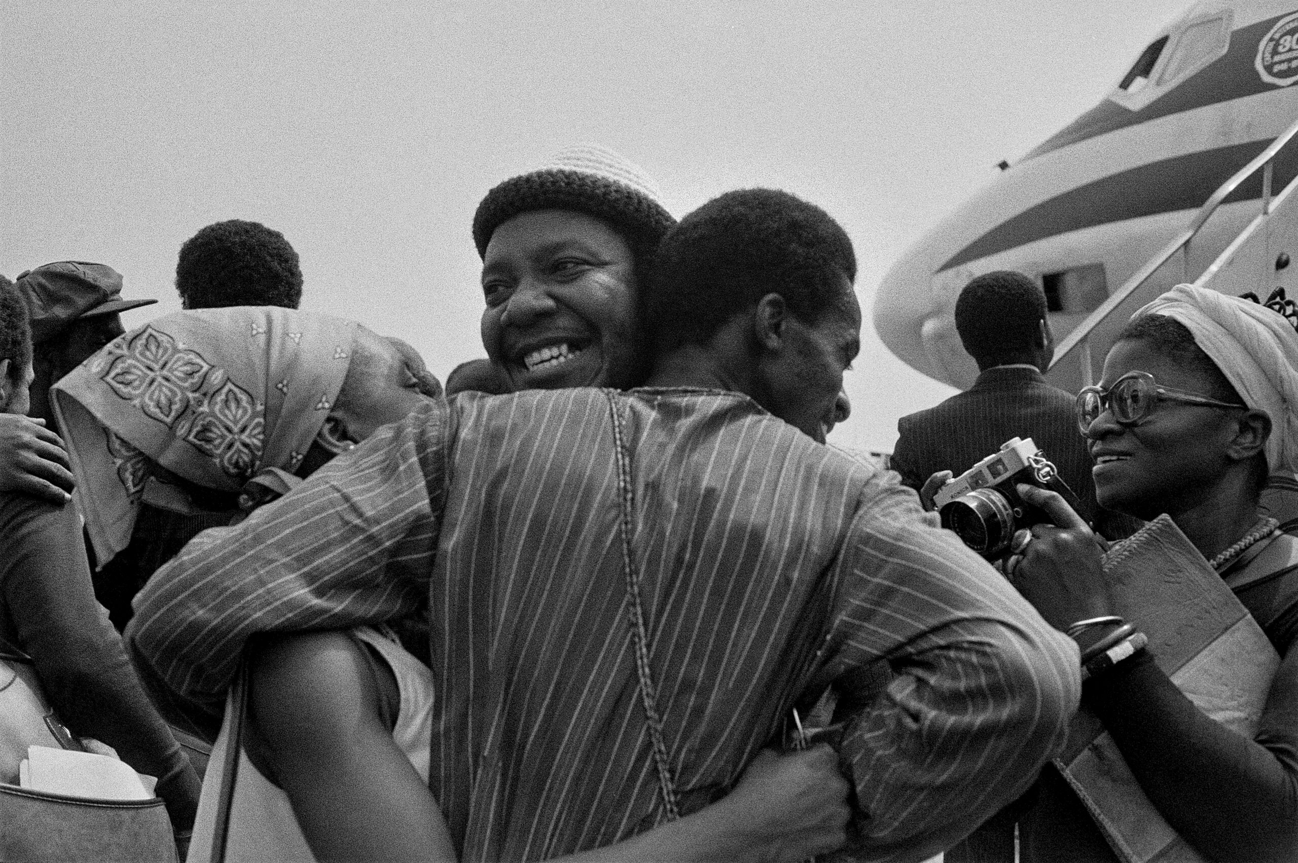 Three people embrace on the tarmac in front of a plane as a photographer positions themself to shoot them close up