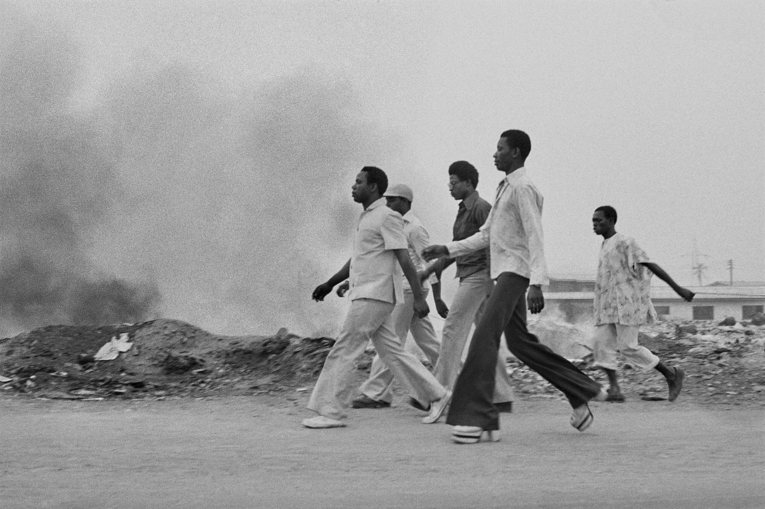Five Black adults walk in unison on a road with a gray cloud in the background
