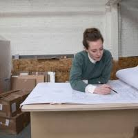 A woman with light skin and shoulder-length brown hair is seen working on large architectural blueprints at a drafting table. She wears a dark green sweater over a white collared shirt. The workspace is industrial, with cardboard boxes and design materials visible in the background.