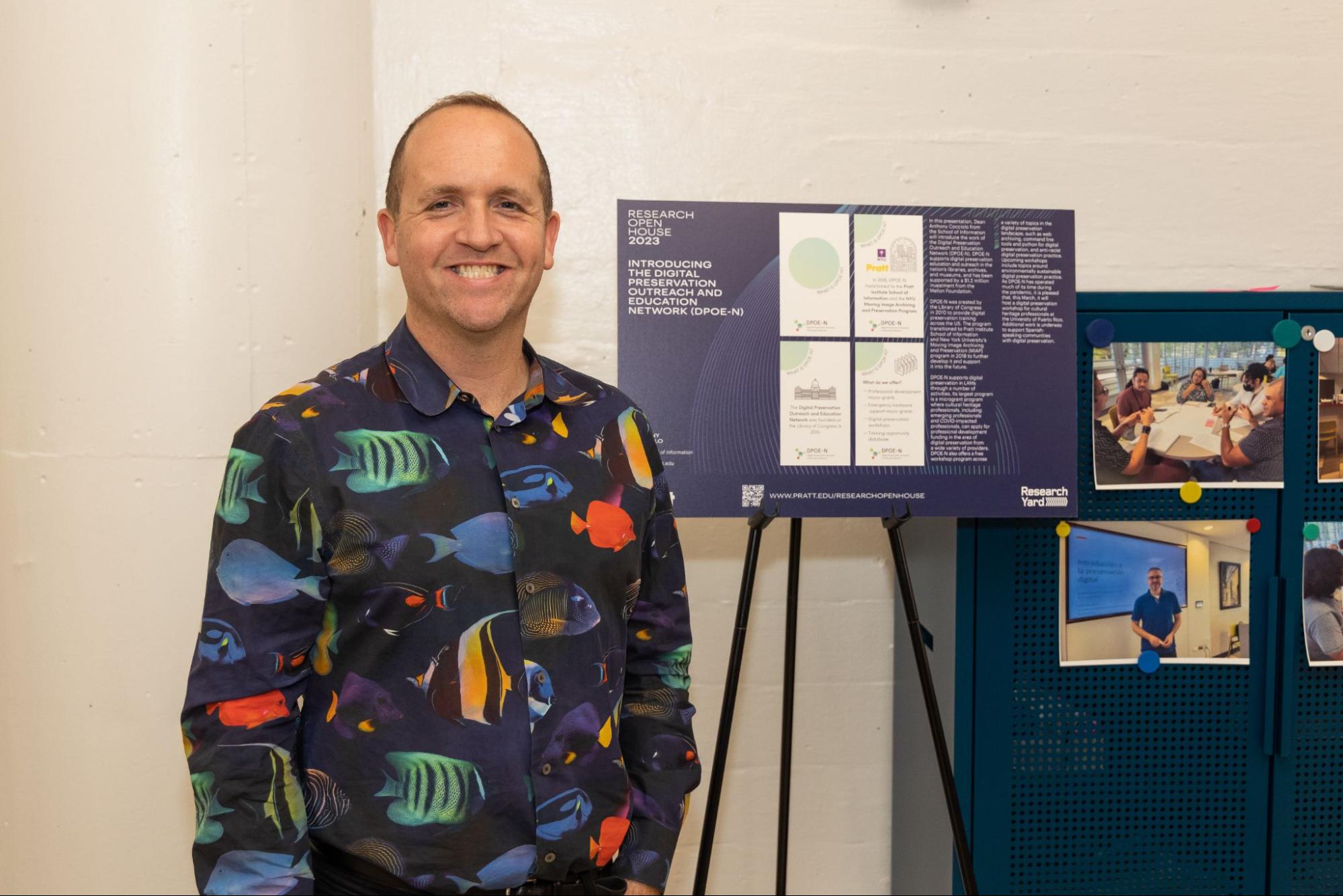 man, standing in front of graphic detailing research open house, smiling, wearing patterned shirt with fish