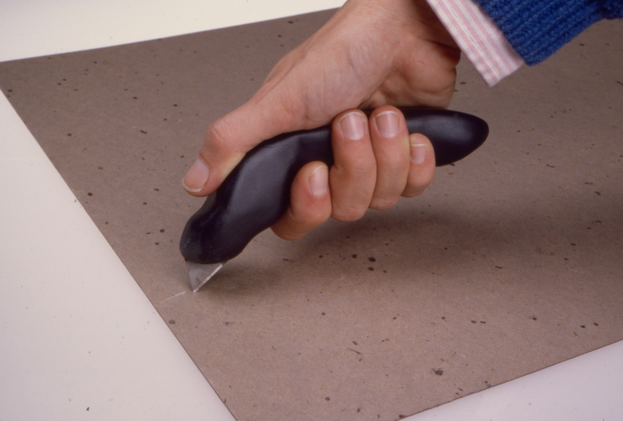 a white hand in a blue sweater slicing a piece of chipboard with a black utility knife