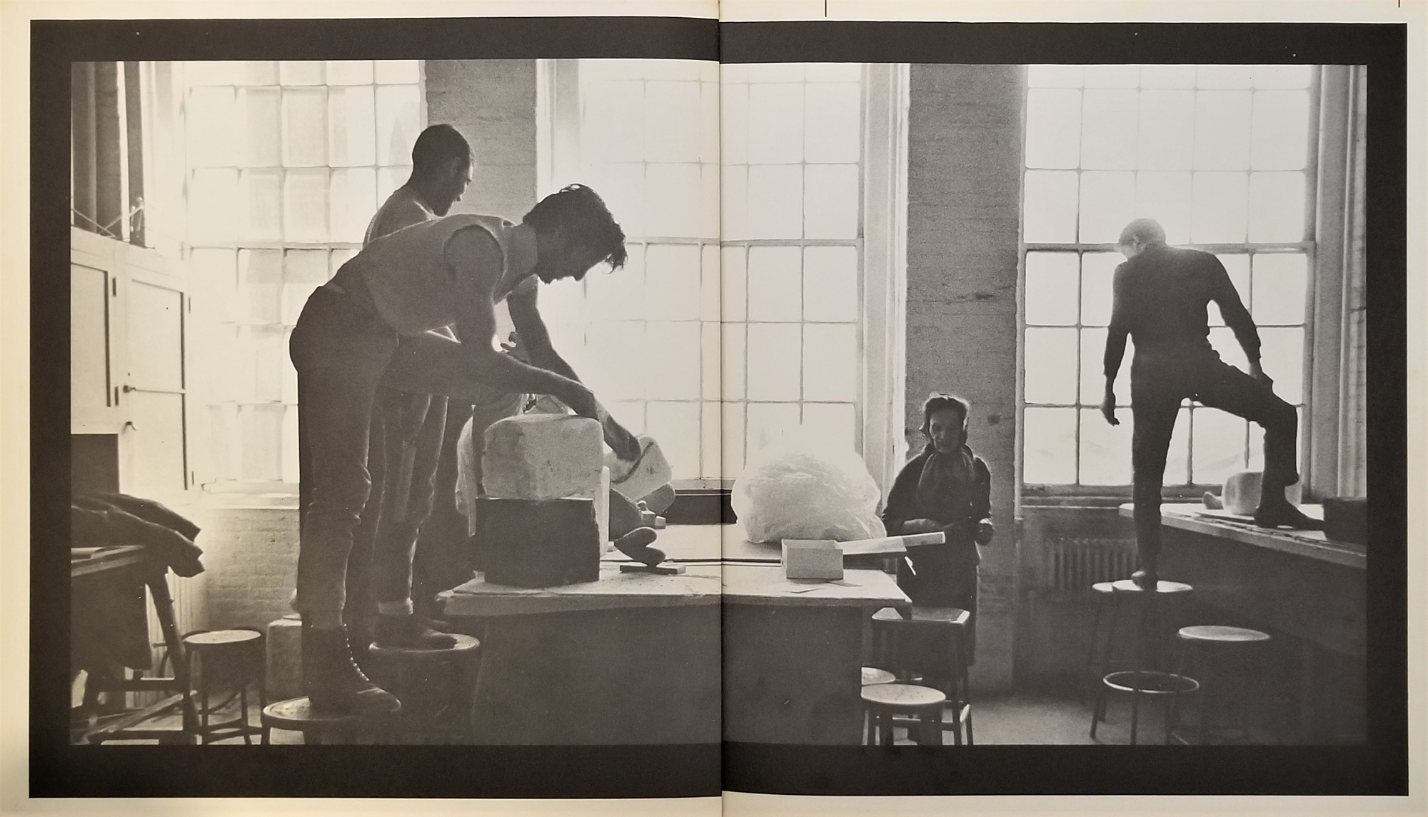 a black and white photograph, taken out of a book, of three young white men in a sculpture studio standing on tables and chairs while an older white woman walks between them on the ground
