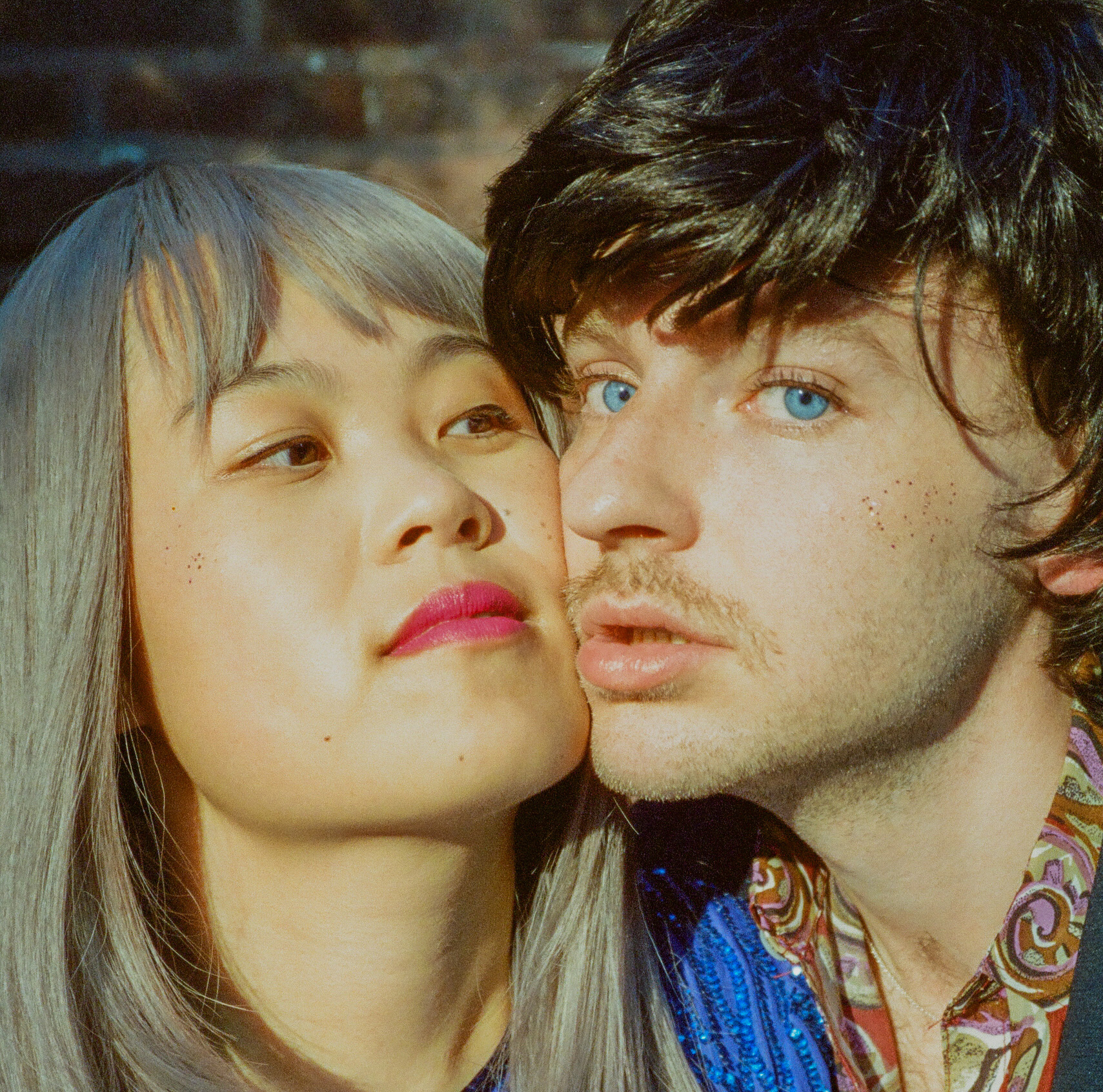 A person wearing lipstick pressing their face against a person with facial hair and blue eyes.