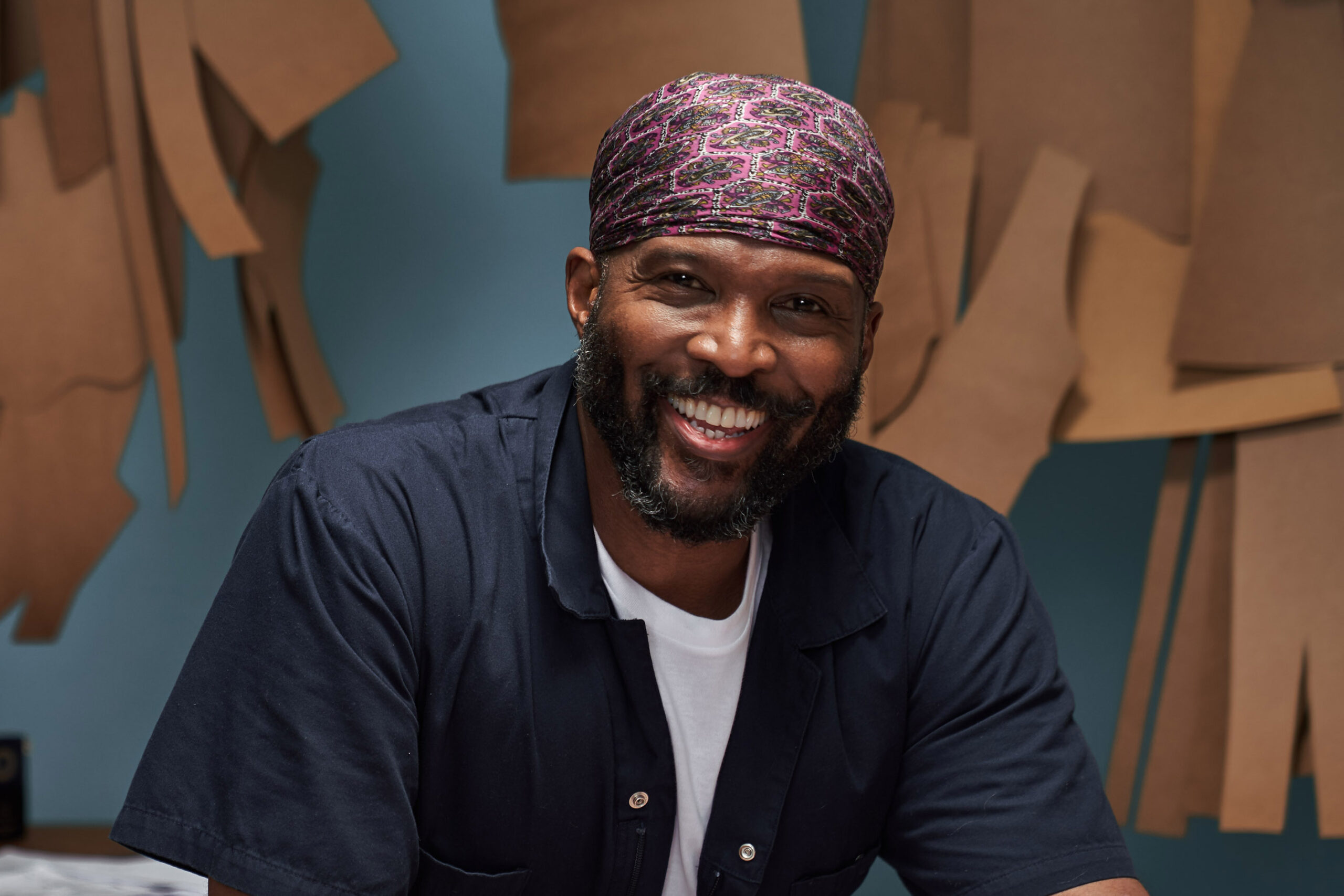 A photograph of Byron Lars smiling, sitting in a studio. He is looking at the camera. Behind him there are brown pieces of paper hanging on the wall.
