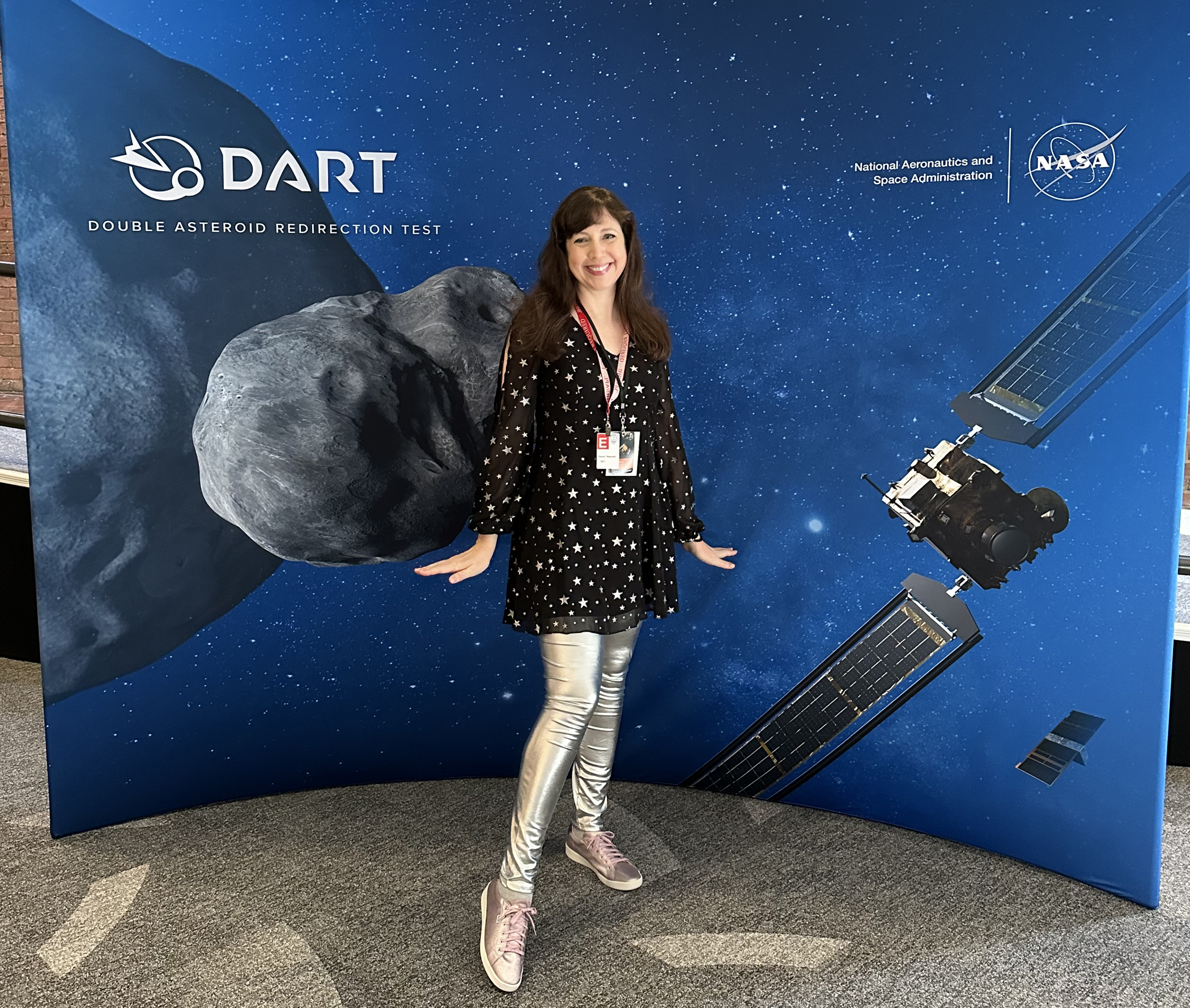 A woman in silver pants and a shirt patterned with silver stars standing in front of a Astrological background designed by NASA.